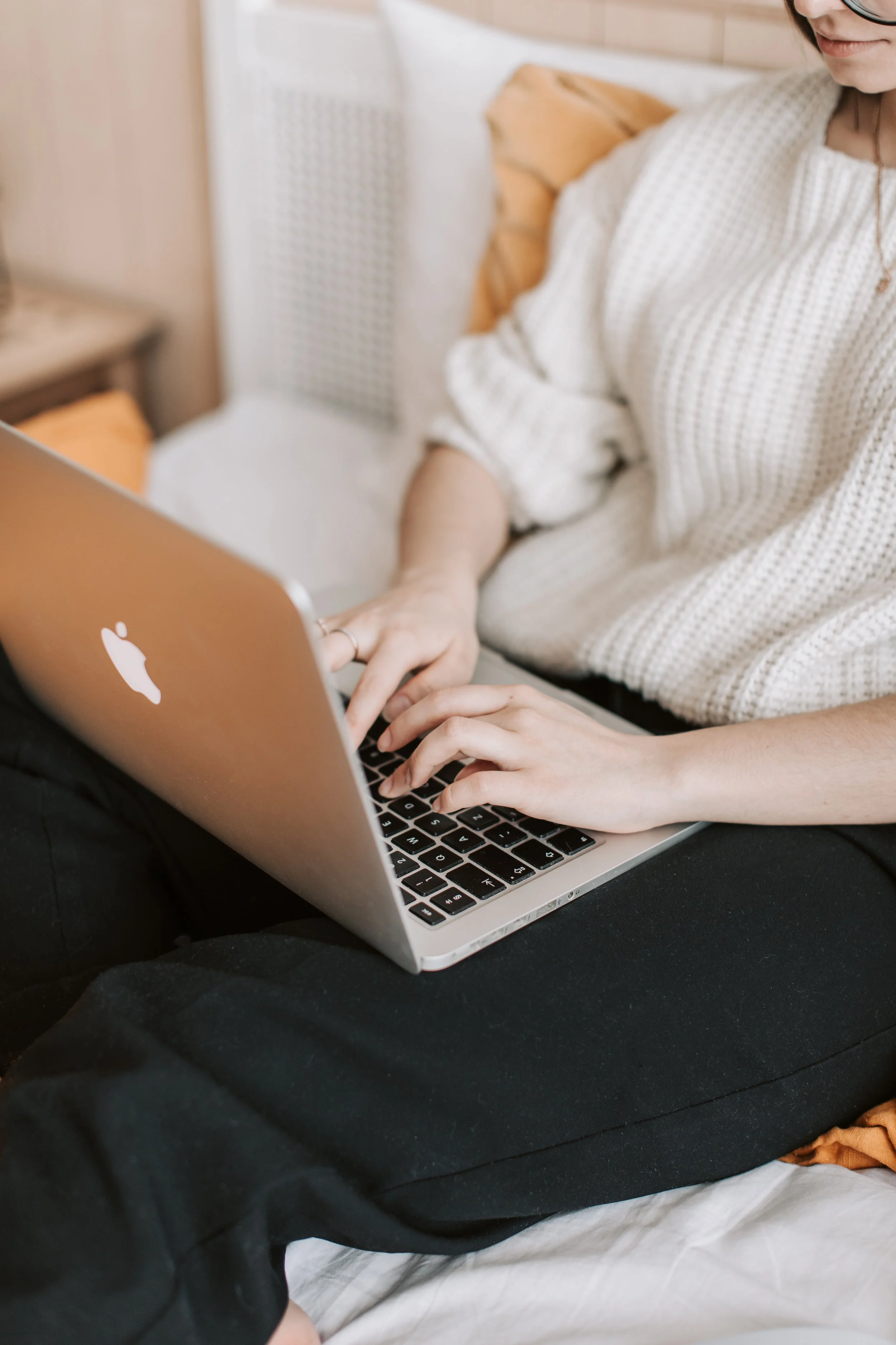 Person sitting on a bed, wearing black pants and a white sweater, using a silver MacBook laptop.