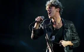 Young man singing into a microphone on stage, wearing a leather jacket and holding a microphone cord.