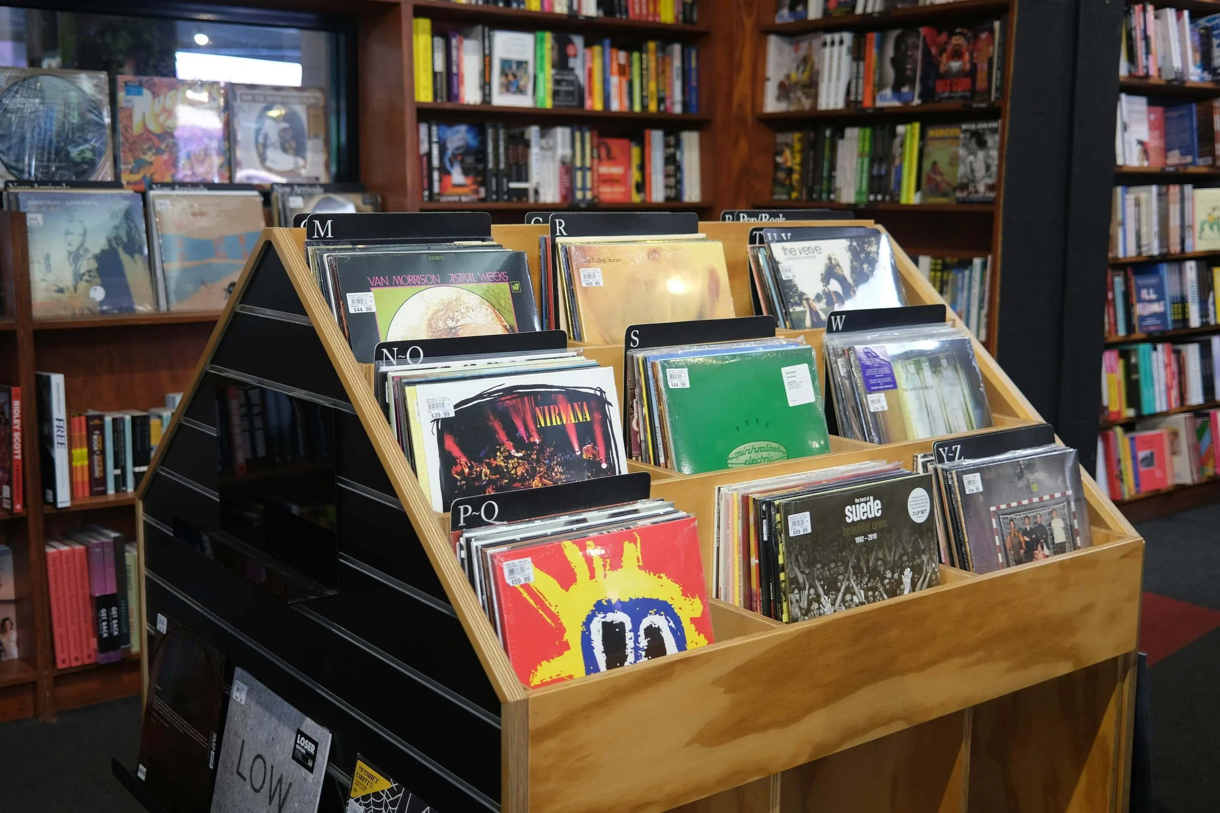 A wooden display rack filled with vinyl records in a bookstore. The records are organized in rows with labeled sections, and bookshelves with various books are visible in the background.