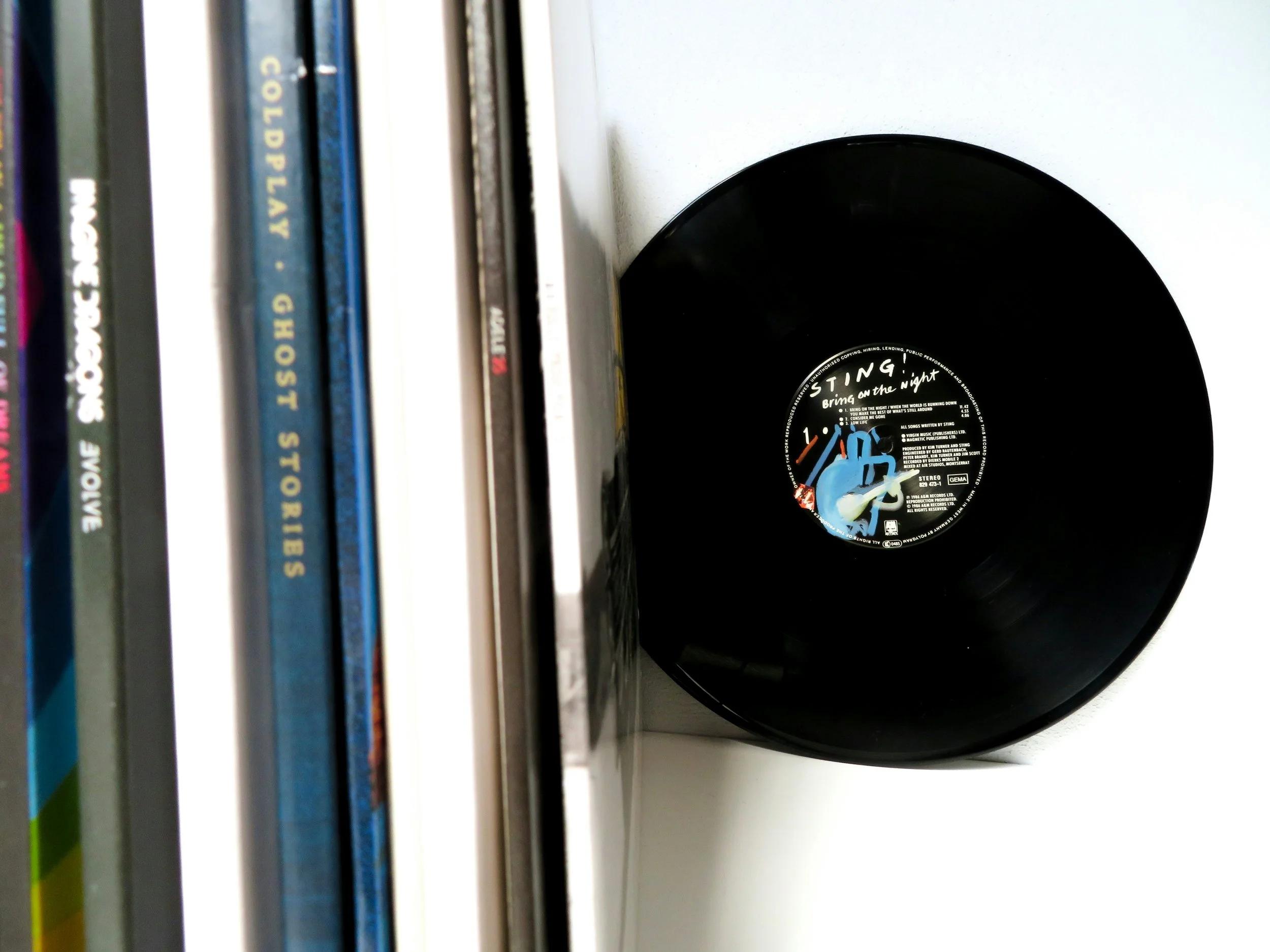 A black vinyl record titled "Bring on the Night" by Sting leaning against a white wall, next to a row of CDs with visible titles such as 'Cold Play,' 'Imara Dunes,' and 'Love'.