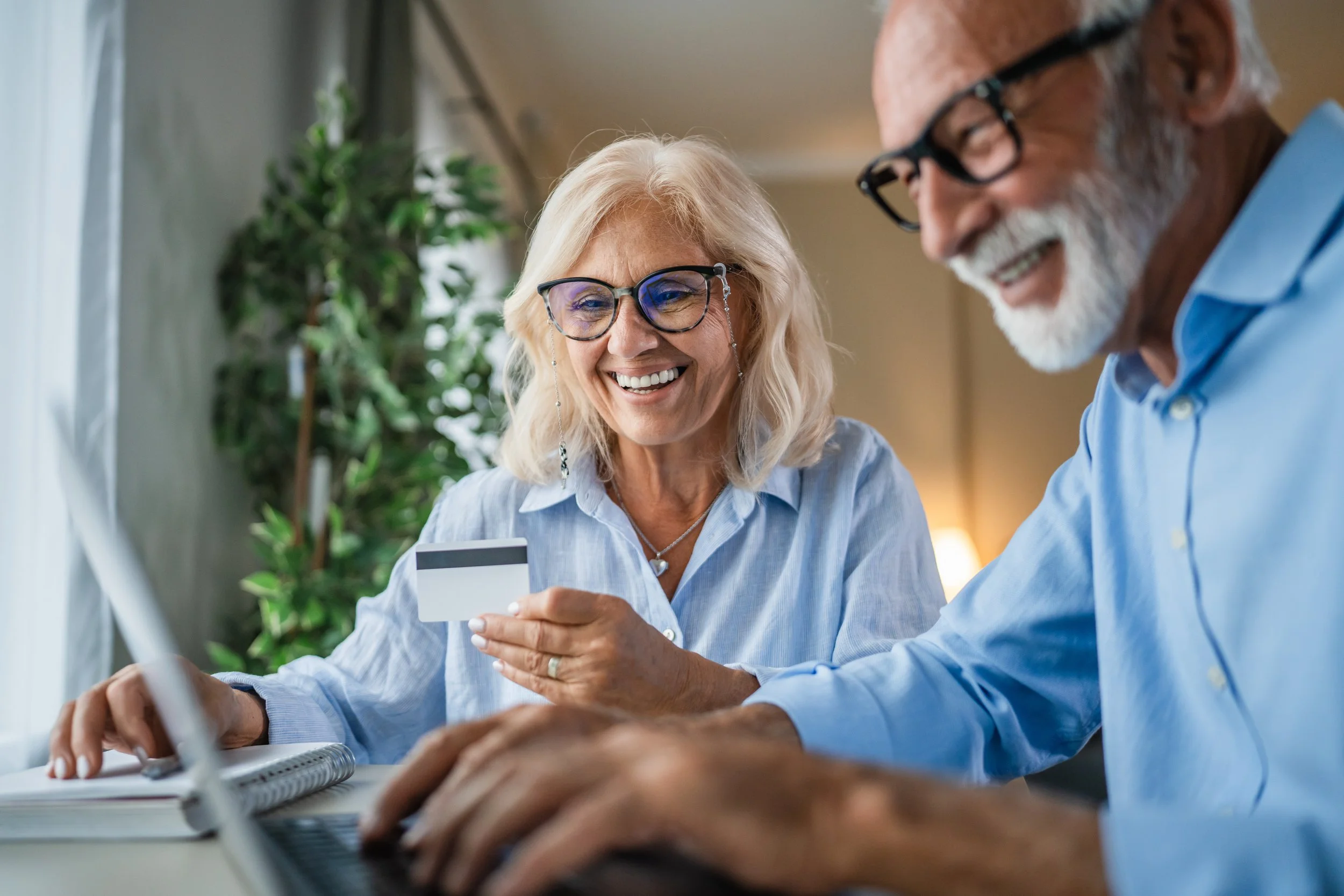 photo of an older couple with glasses smiling at a laptop with a credit card in hand.
