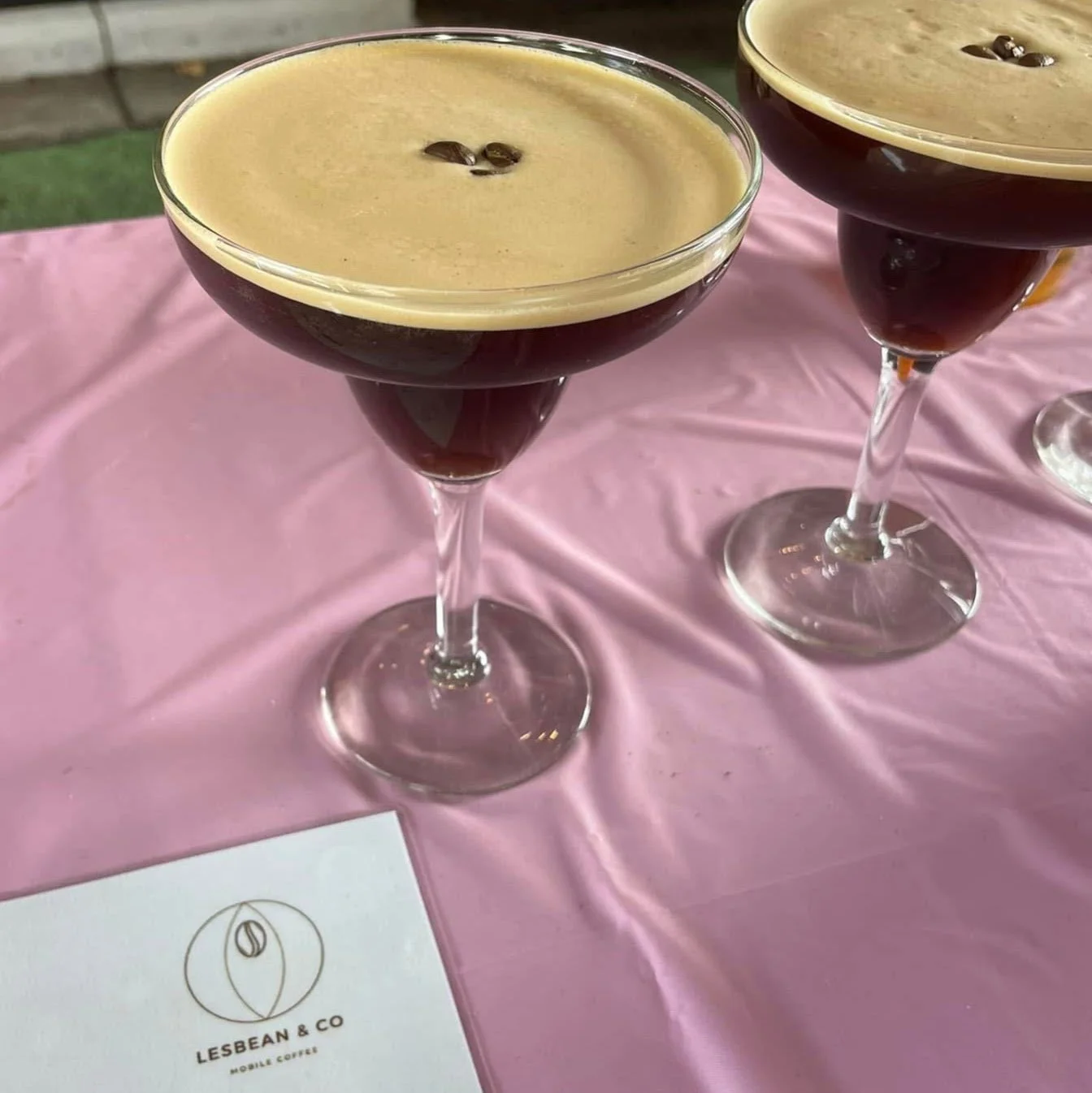 Two espresso martini cocktails in coupe glasses with dark liquid and a creamy foam top, garnished with coffee beans, on a pink tablecloth with a Lesbean & Co card.