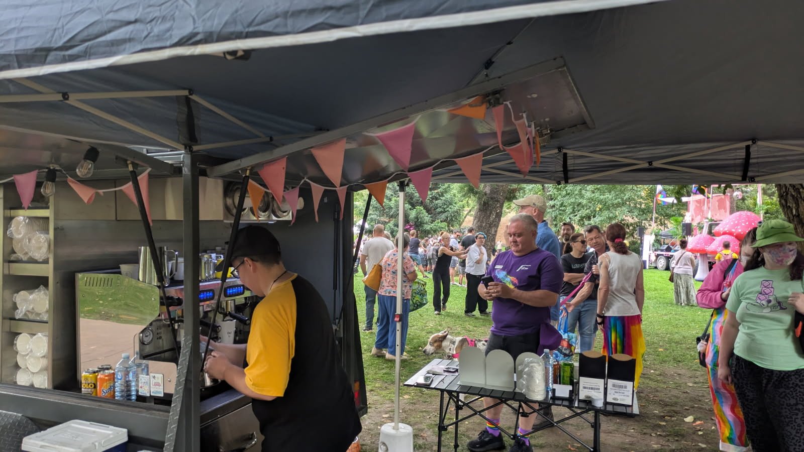 A coffee van at Bendigo Pride Party in the Park with people in line and others walking in a park, with bunting in the Lesbian flag colours hanging from the cart's canopy.