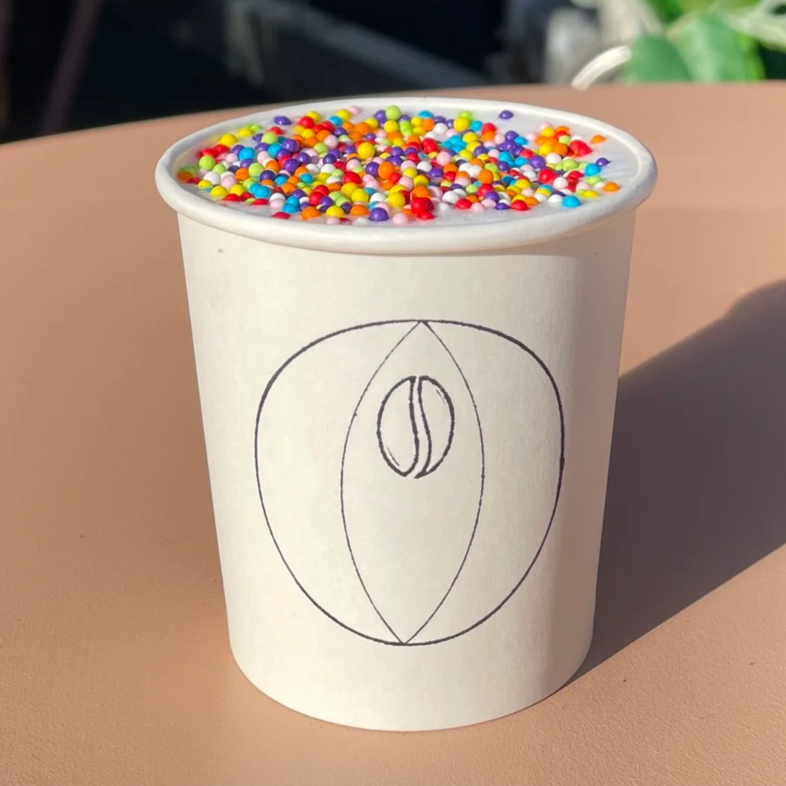 A small white paper cup filled with frothed milk topped with rainbow sprinkles. The Lesbean & Co logo is stamped on the front of the cup.