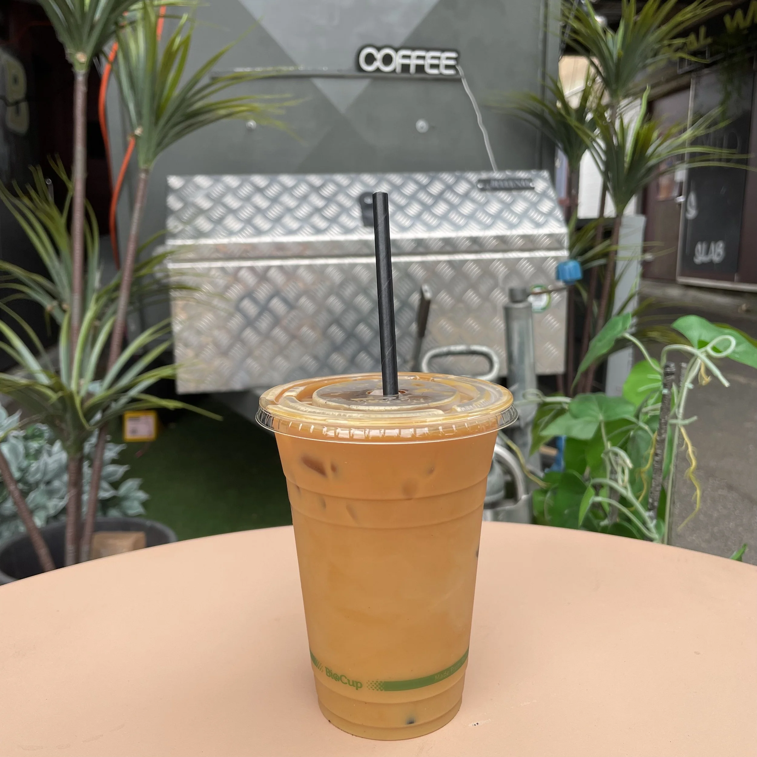 A cold iced coffee in a clear plastic cup with a black straw, placed on a pink tabletop outdoors, with green plants and a coffee van in the background.