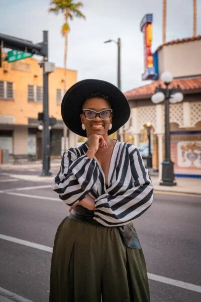 A woman smiling and posing on a city street, wearing a black wide-brimmed hat, glasses, a striped blouse, and olive-green pants.