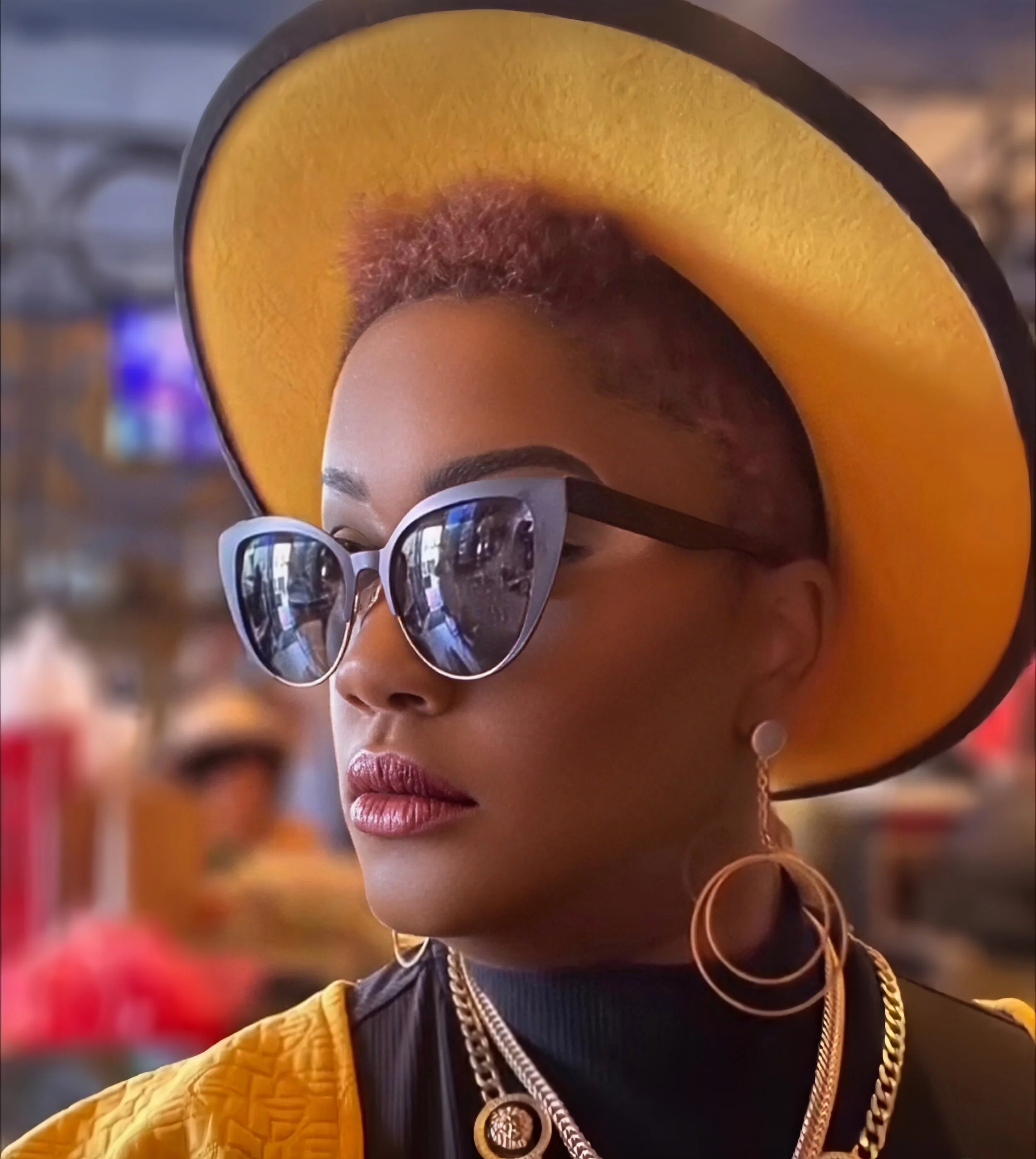 A woman wearing large sunglasses and gold jewelry, with a wide-brimmed hat shaped like a slice of peach, poses confidently in an indoor setting.