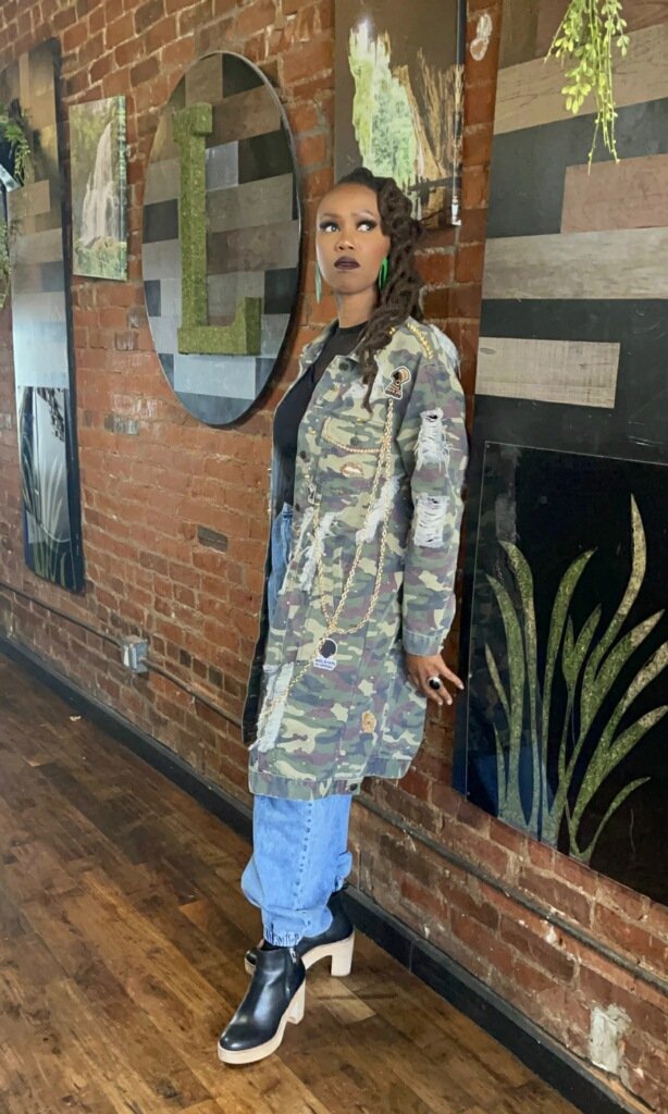 A woman with dark lipstick, green earrings, and styled hair standing indoors against a brick wall and artwork. She is wearing a camouflage coat with patches, ripped jeans, and unique black and white chunky heeled shoes.