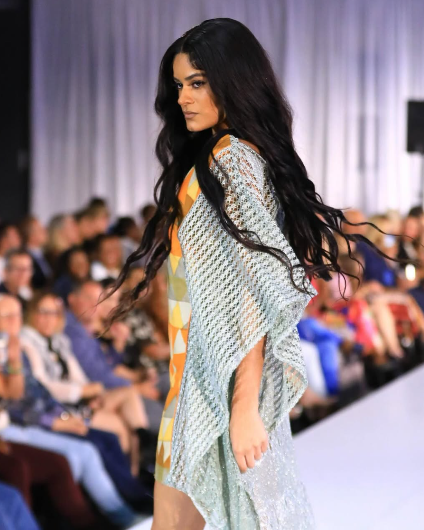 Model walking on runway wearing shiny, textured metallic dress with colorful geometric design on sleeve, with seated audience in background