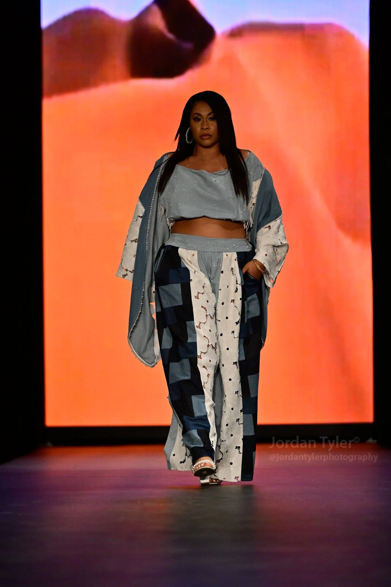 A woman walking on a runway wearing a blue and white patchwork outfit, with an abstract orange background.