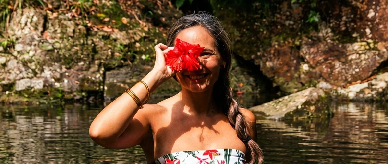 A woman smiling and holding a red flower to her eye, standing by a river with rocks and trees in the background.
