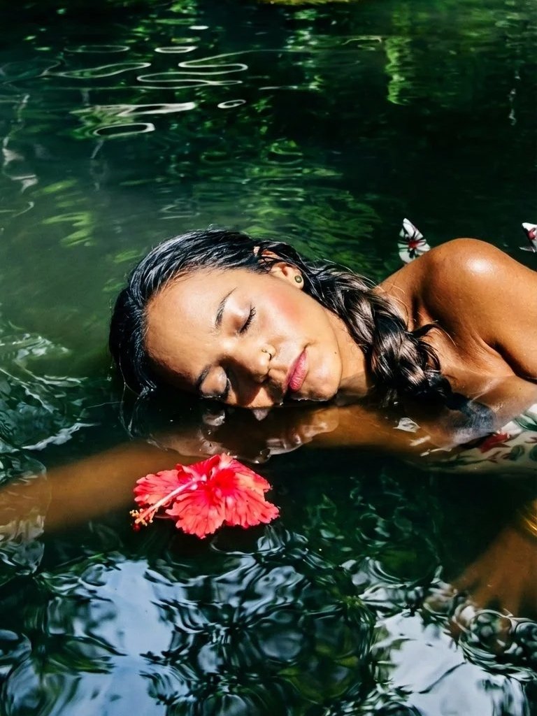 A woman calmly floating in a body of water with her eyes closed, a red hibiscus flower near her face, surrounded by greenery reflected in the water.
