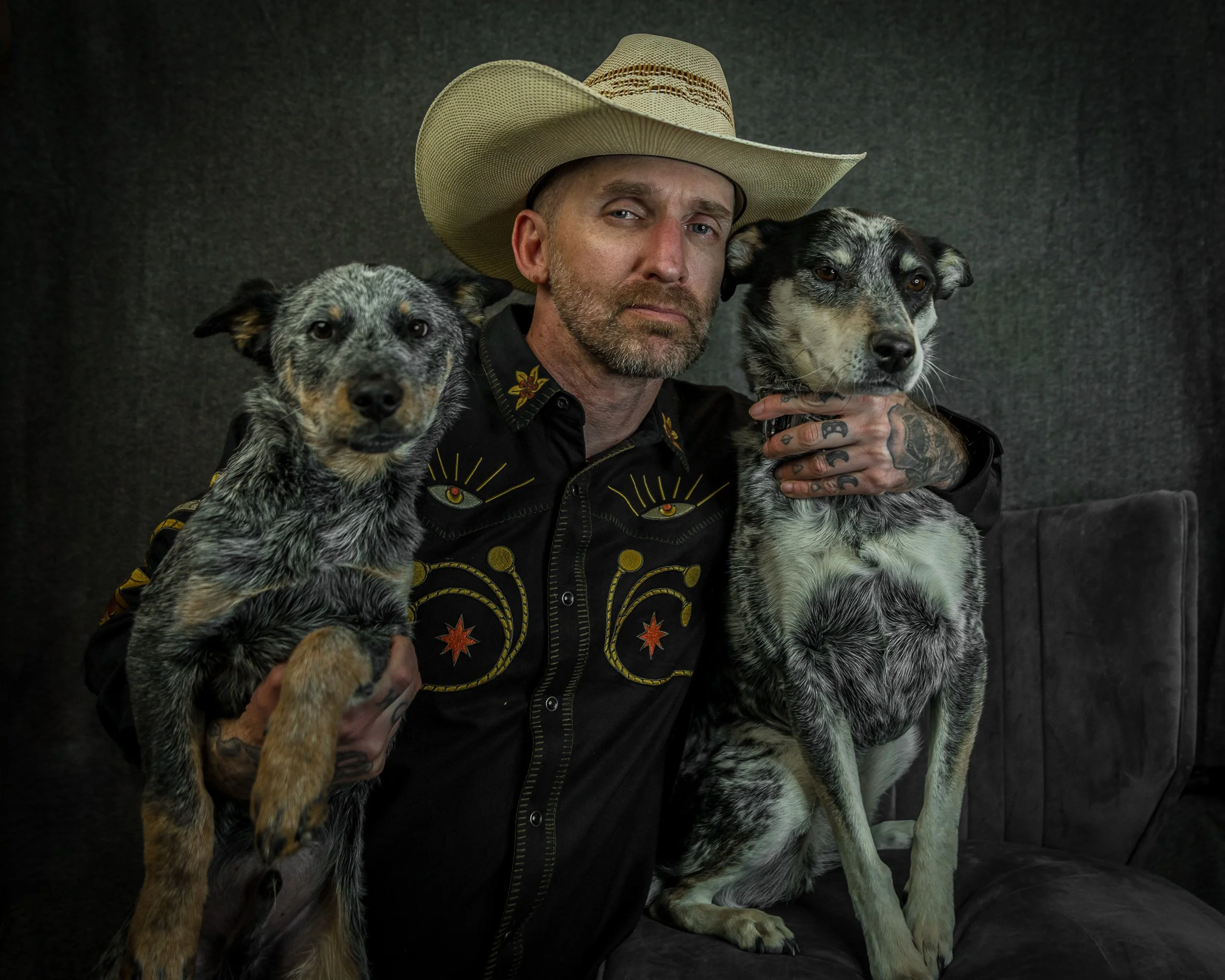A man wearing a cowboy hat and a black shirt with decorative embroidery holds two dogs with black, white, and gray patches against a dark background.