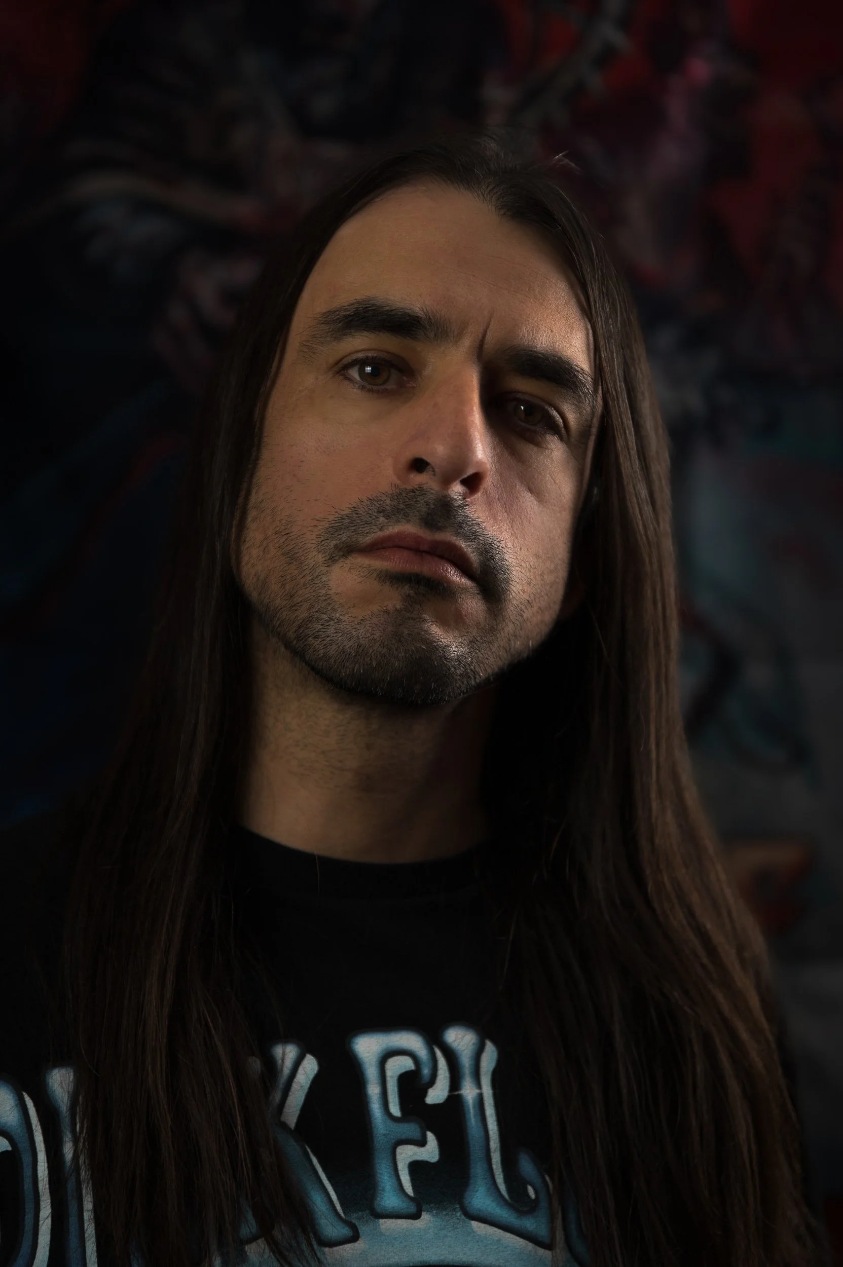 A person with long brown hair, facial hair, and a serious expression, wearing a black shirt with blue lettering, looking directly at the camera.