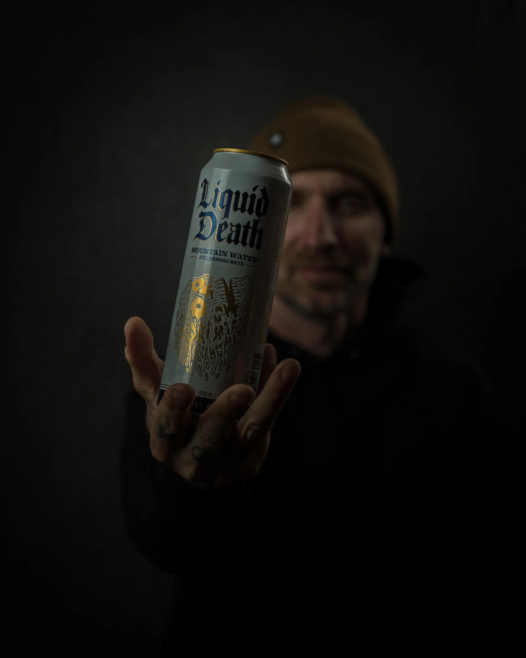 A man wearing a brown beanie and glasses holding a can of Liquid Death Mountain Water towards the camera, with a dark background.