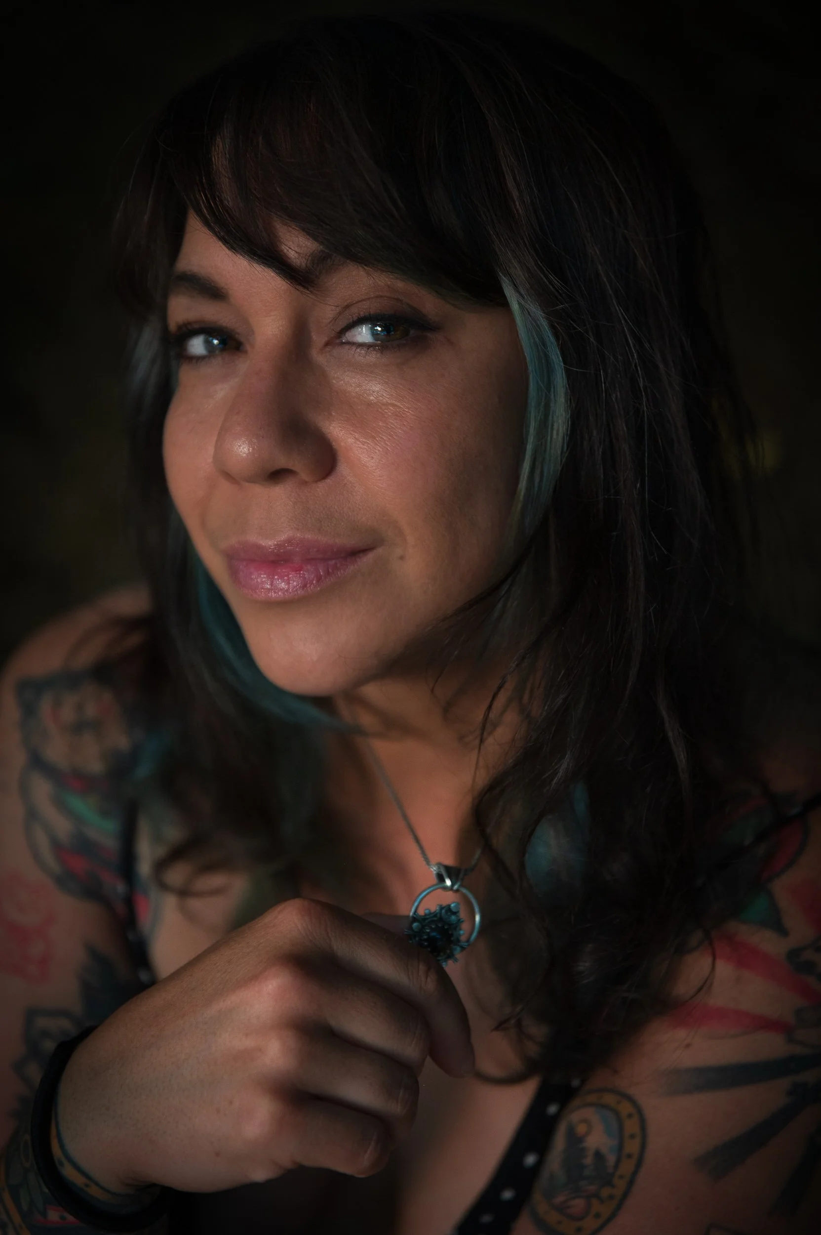 A woman with dark hair and tattoos on her arms, wearing a black polka-dot top, is holding a small object in her hand. She has blue highlights in her hair, and blue eyes, and is wearing a necklace with a circular pendant.