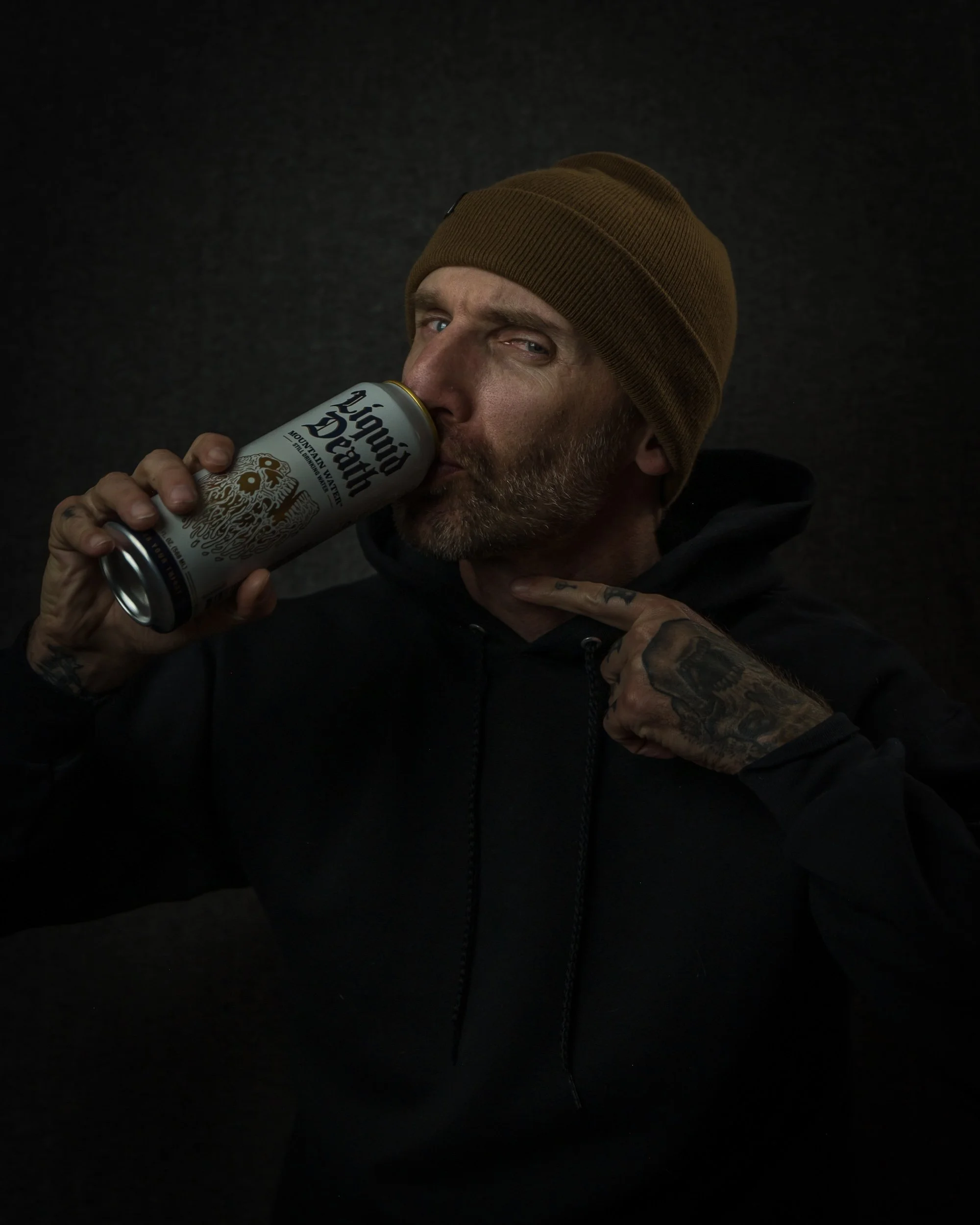 A man with a beard and tattoos on his hand and neck, wearing a brown beanie and black hoodie, drinks from a white can of Mountain Valley Sparkling Water against a dark background.
