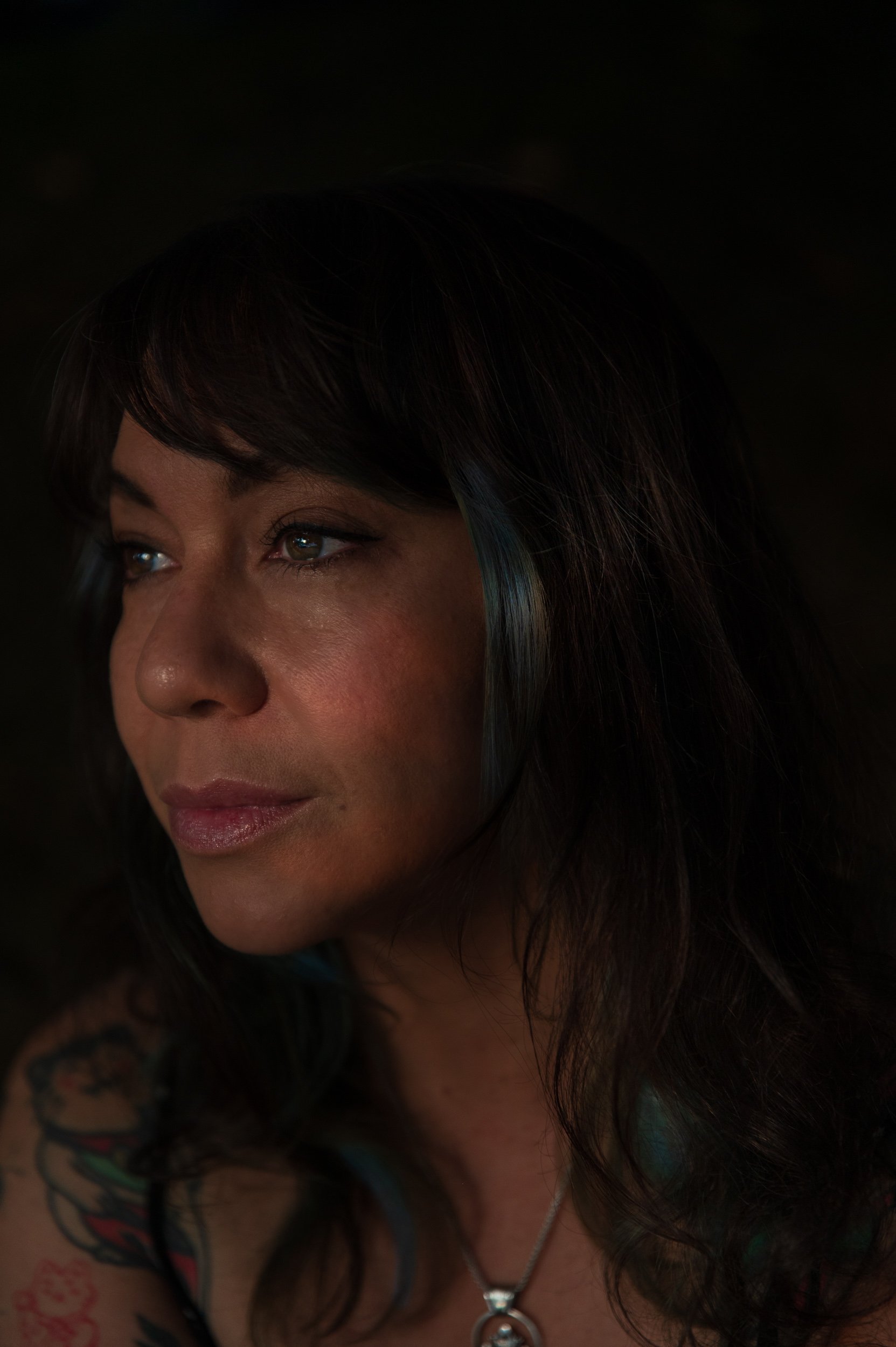 Close-up of a woman with dark hair, wearing makeup, a necklace, and tattoos on her arm.