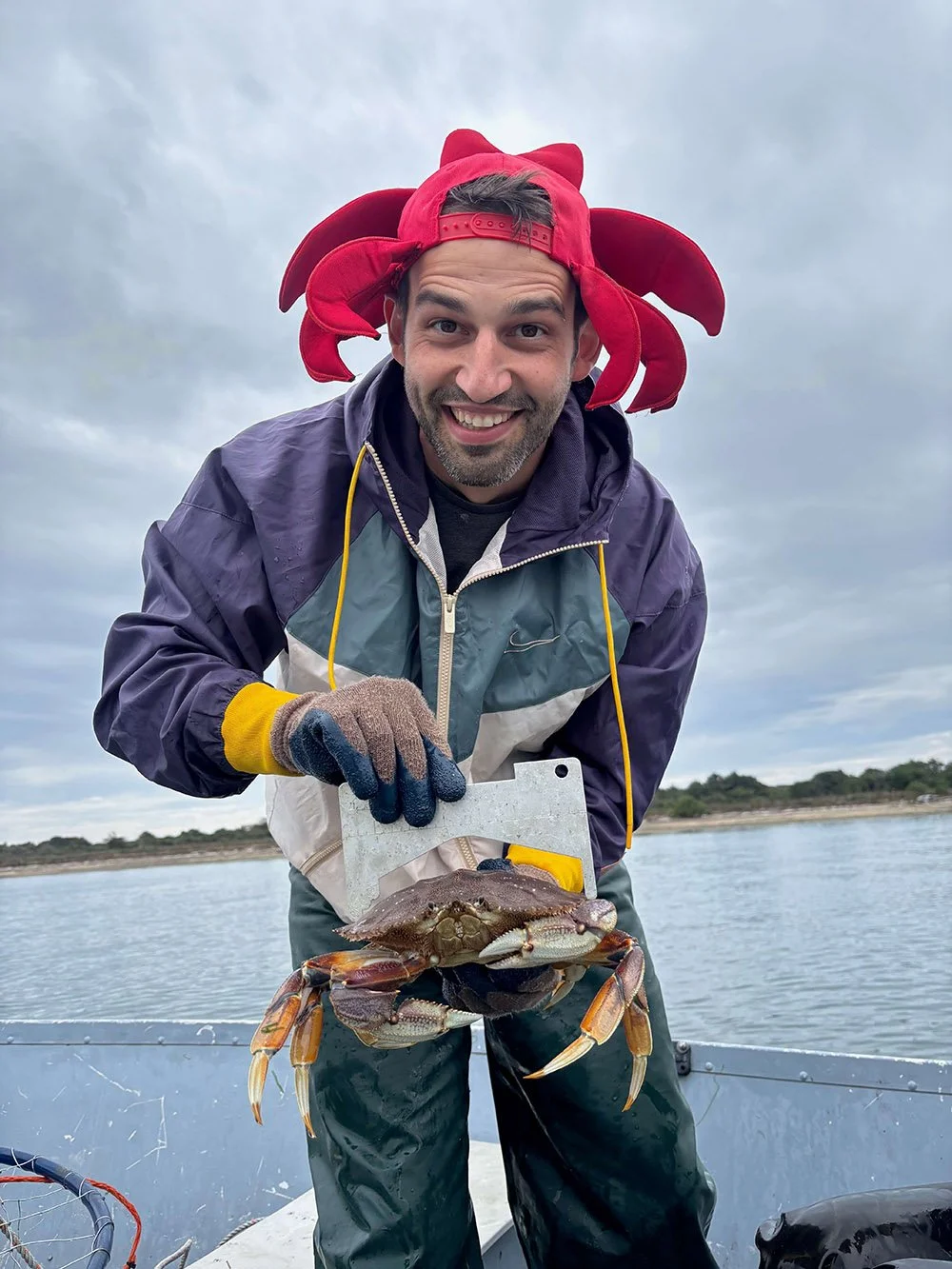 Man holding a crab on a boat, wearing a hat with red octopus tentacles, waterproof jacket, and gloves, with a cloudy sky in the background.