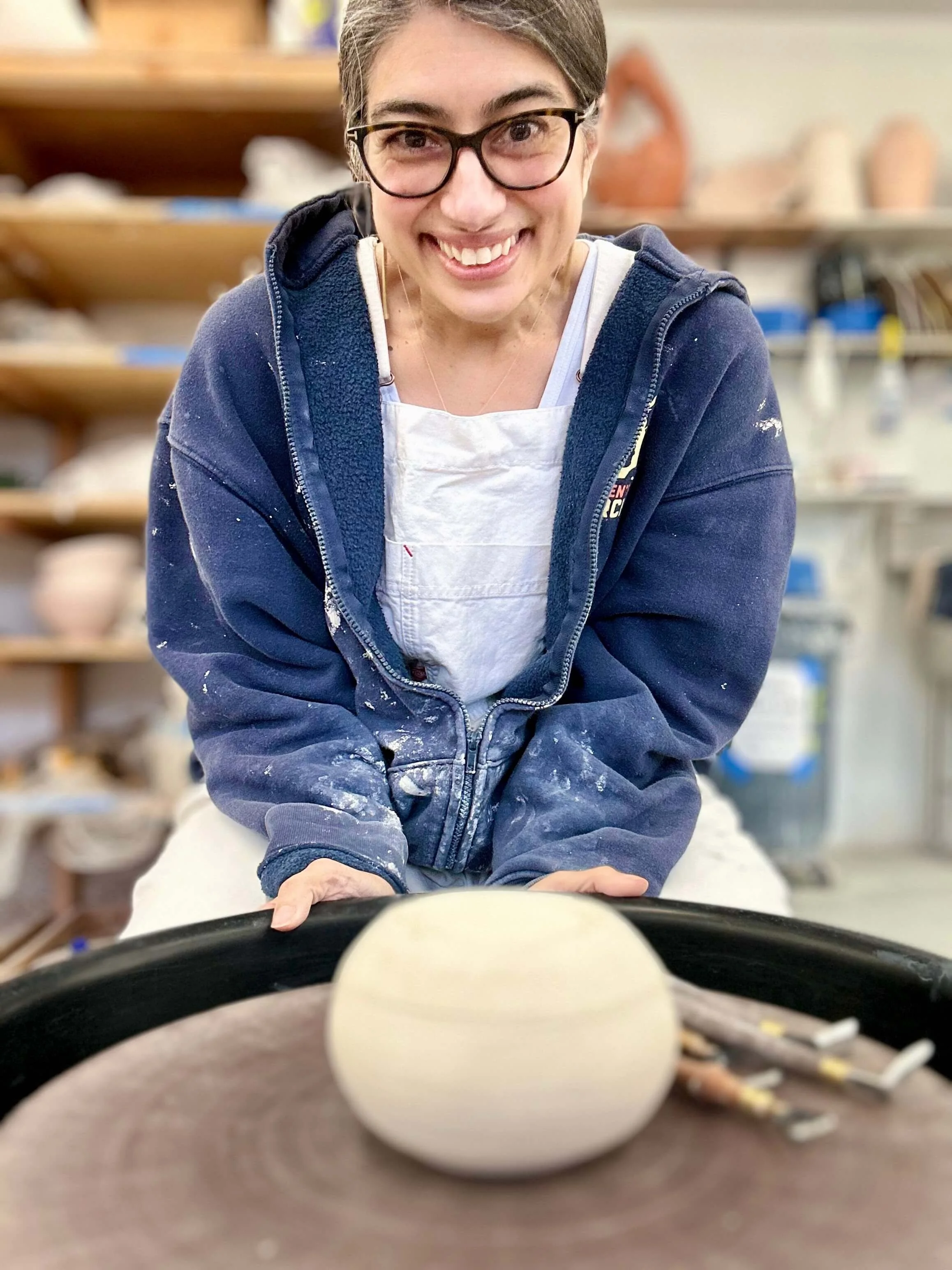 A woman shaping a ceramic bowl on a pottery wheel in a pottery studio, smiling at the camera.