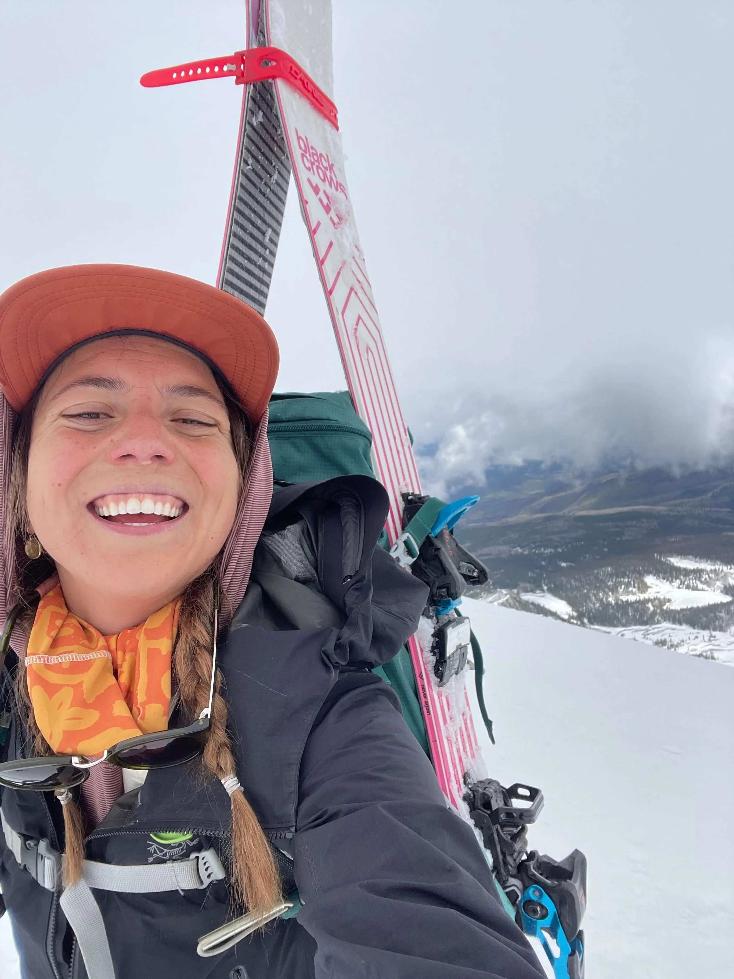 Smiling woman wearing an orange cap and orange patterned scarf, taking a selfie on a snowy mountain with ski gear and a pink and white snowboard in the background.