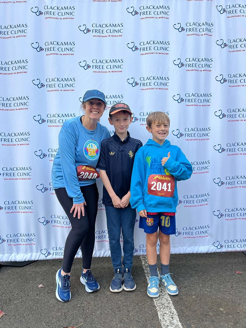 A woman and two boys standing in front of a backdrop with the logo of Clackamas Free Clinic, participating in a 5K race event. The woman is wearing a blue shirt, black leggings, a blue cap, and running shoes, with a race bib numbered 2040. One boy is wearing a black jacket, blue jeans, and a cap, while the other boy is in a blue hoodie, shorts, and sneakers, giving a thumbs up.