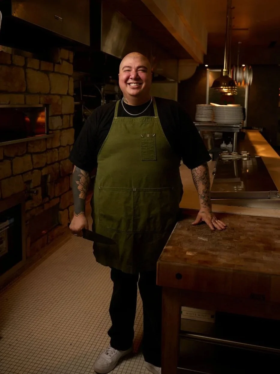 Line cook at Herb & Sea smiling while standing in a kitchen holding a large fish hanging upside down.