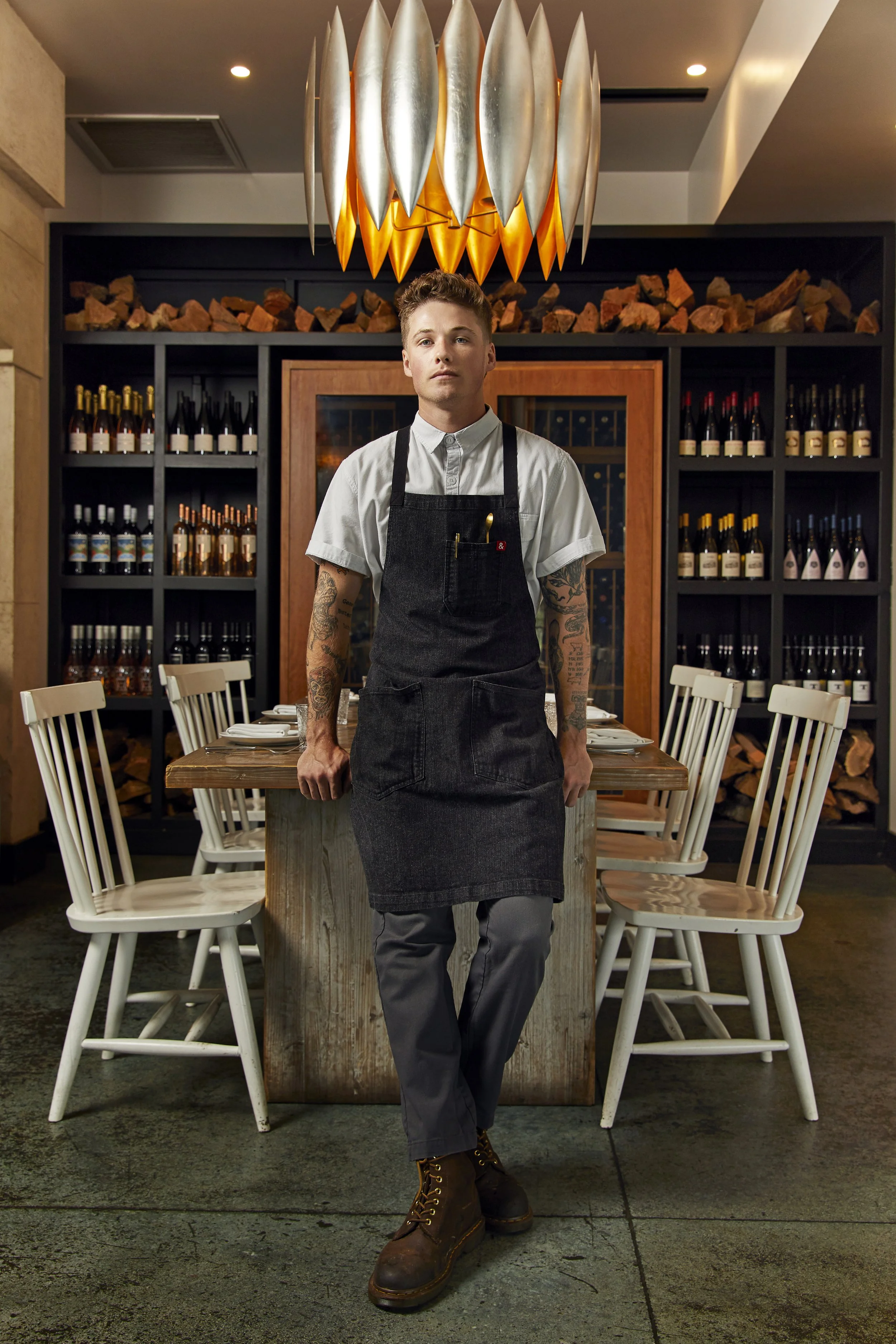 Chef Aidan Owens in a dark apron standing in the dining room at Herb & Sea, Encinitas CA — wine shelves and firewood stacked behind him