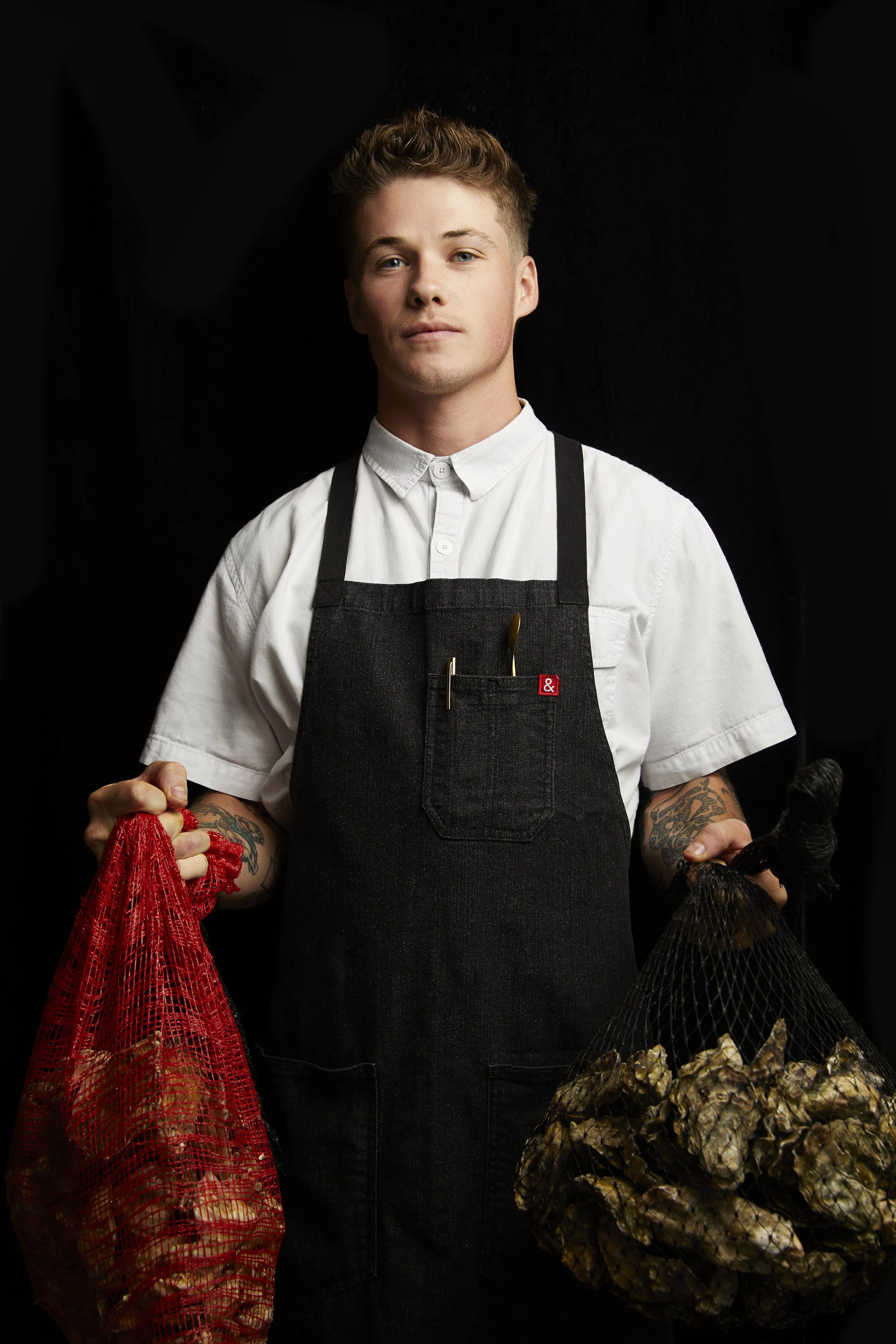 Culinary Director Aidan Owens in a dark apron and white chef coat, holding two bags of raw oysters