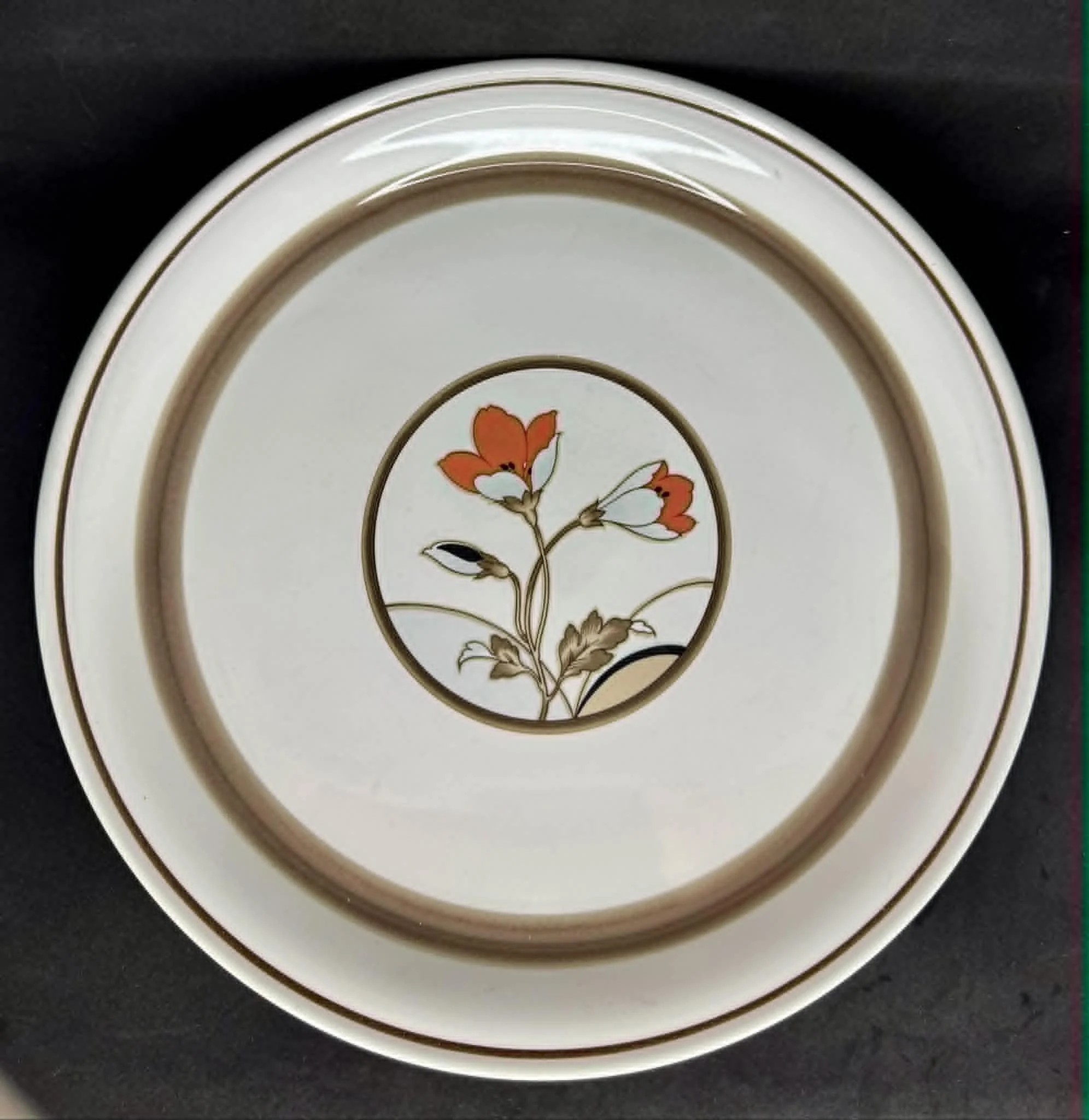 A white ceramic plate with thin brown lines on the edges, featuring floral decoration in the center, resting on a black surface.