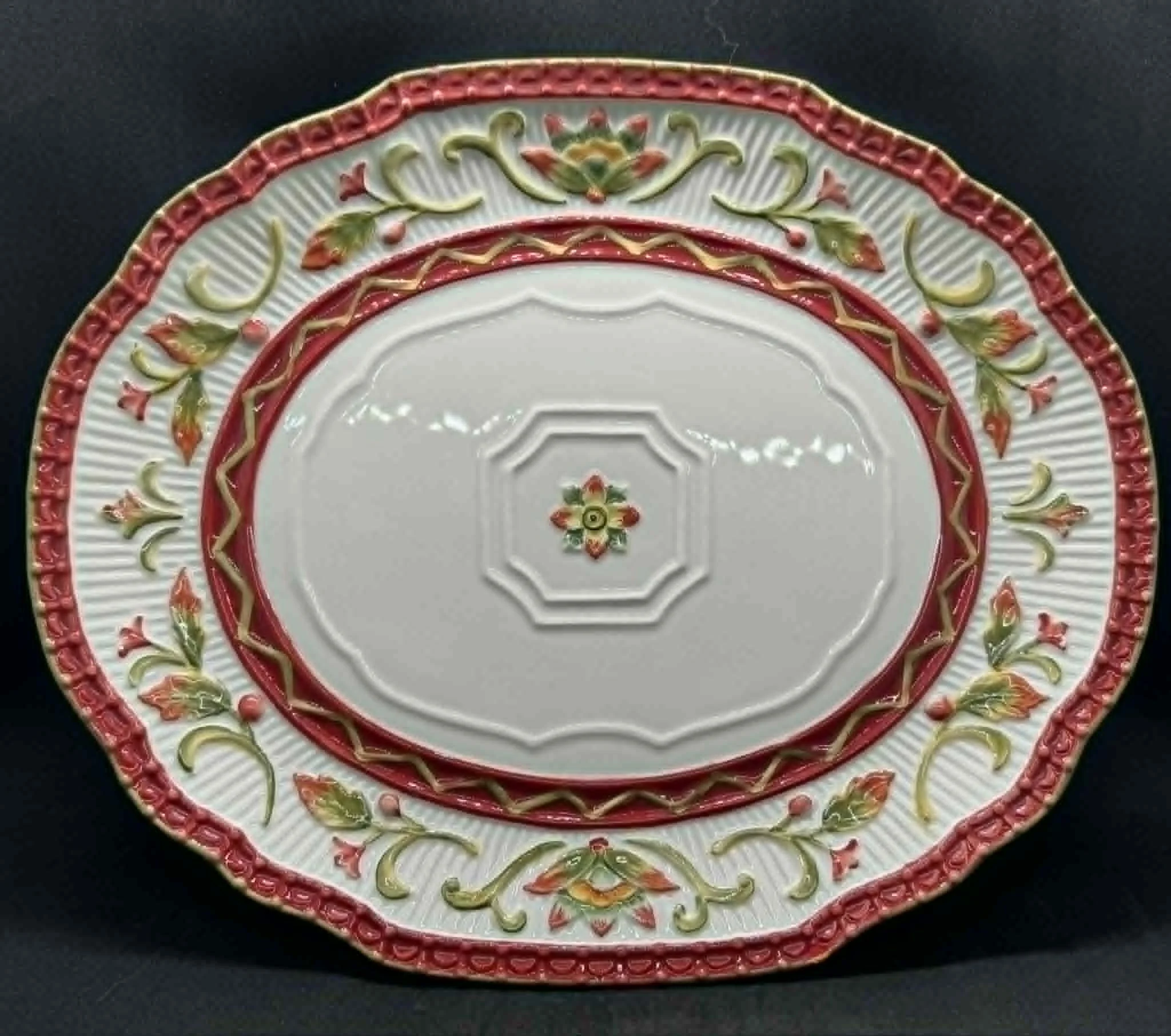 Decorative ceramic plate with intricate floral patterns in red, green, and gold, featuring a multi-layered rim, placed on a dark surface.