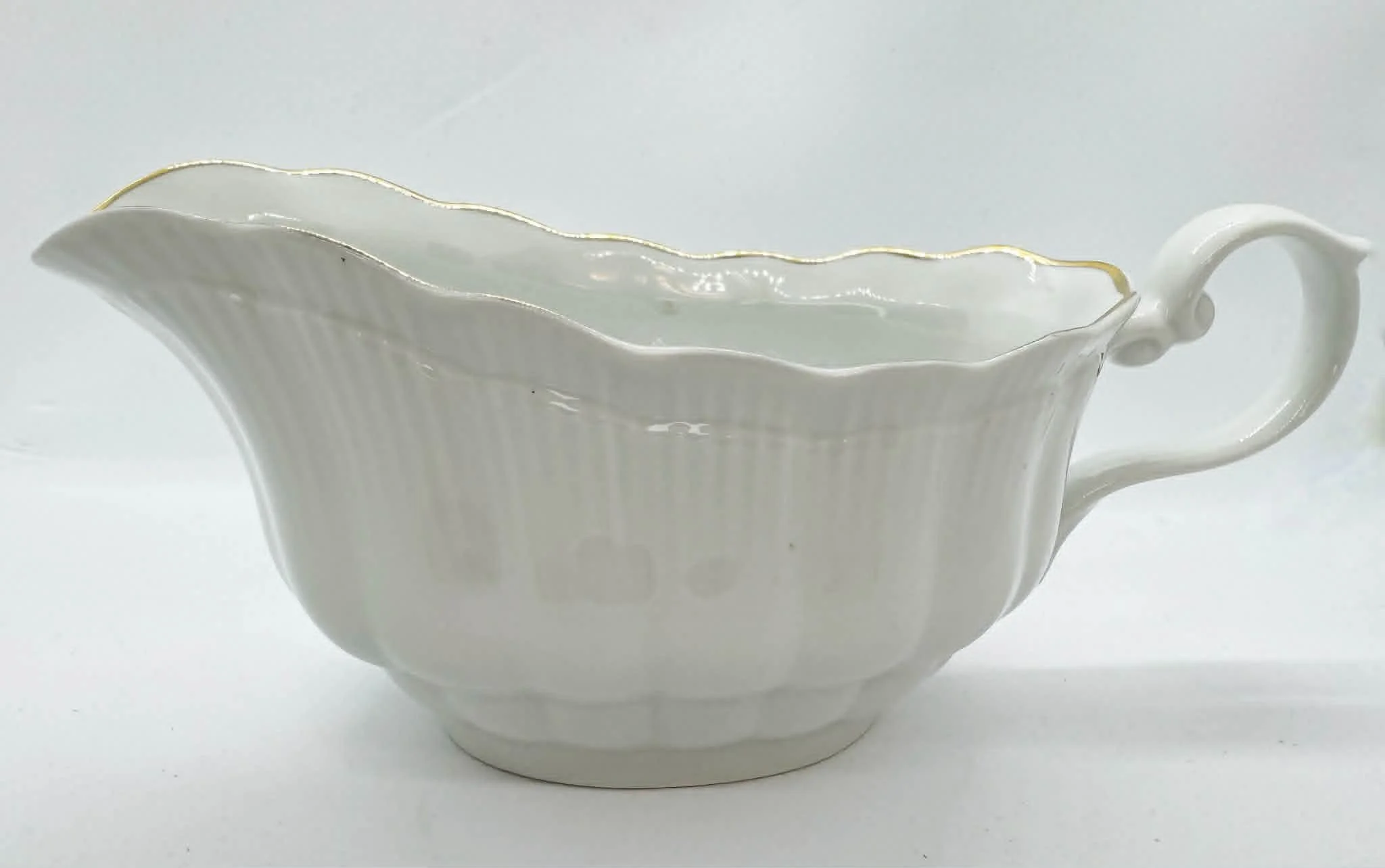 A white porcelain creamer with a gold trim, placed on a white surface against a plain background.