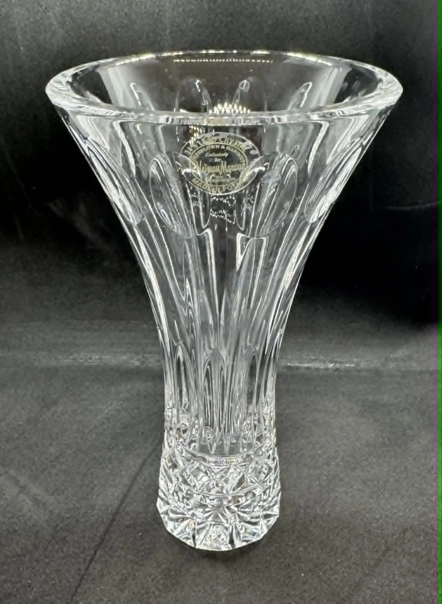 A clear, cut-glass vase with intricate patterns and a gold sticker on the front, placed on a dark surface.