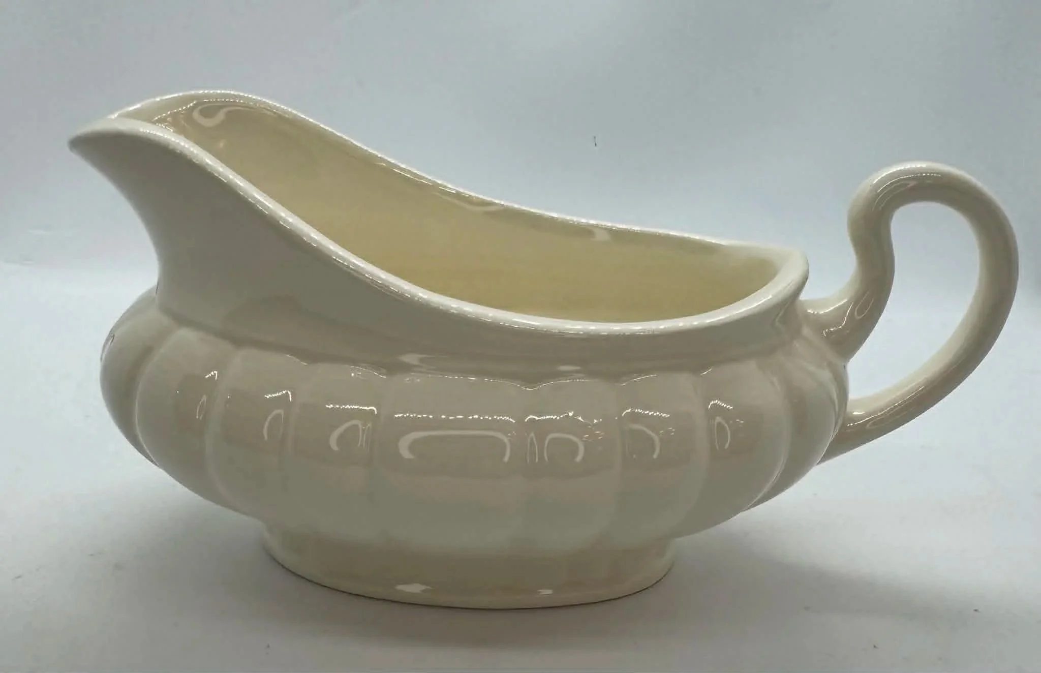 Cream-colored ceramic gravy boat with a curved handle and decorative pattern around its side, placed on a white surface against a gray background.