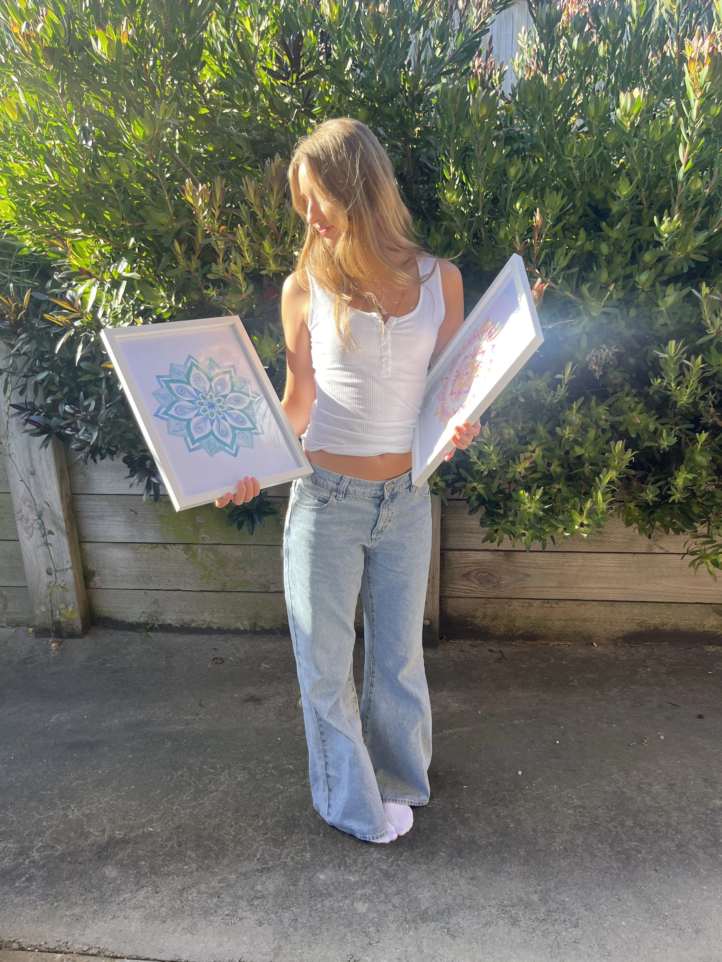 A young woman with light brown hair wearing a white sleeveless top and light blue jeans stands outdoors holding two framed art prints featuring colorful mandala designs.