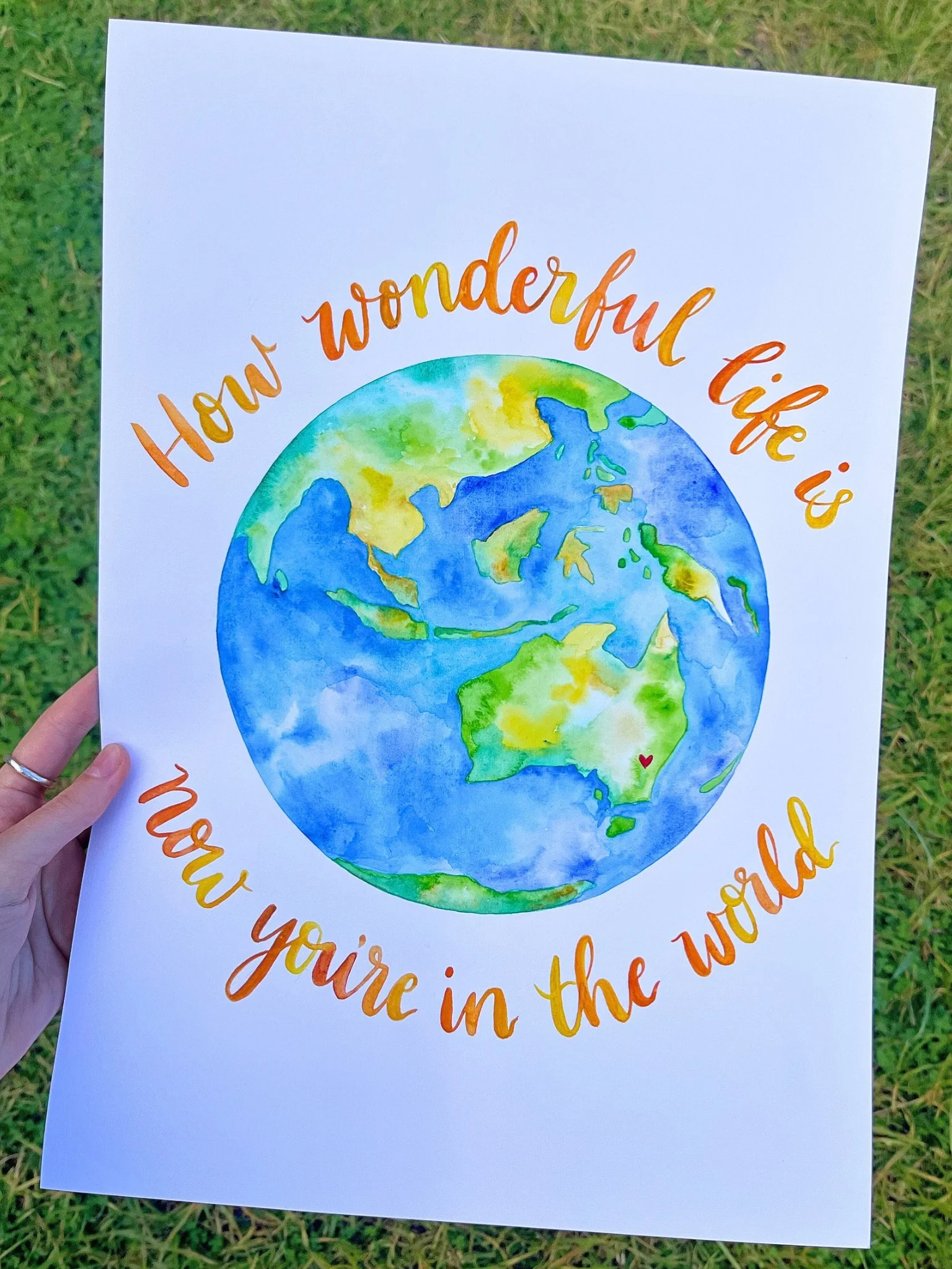 Watercolor illustration of Earth with the quote 'How wonderful life is when you're in the world' surrounding it. The Earth is depicted with blue oceans, green, yellow, and brown land masses, and a small red heart on Australia.