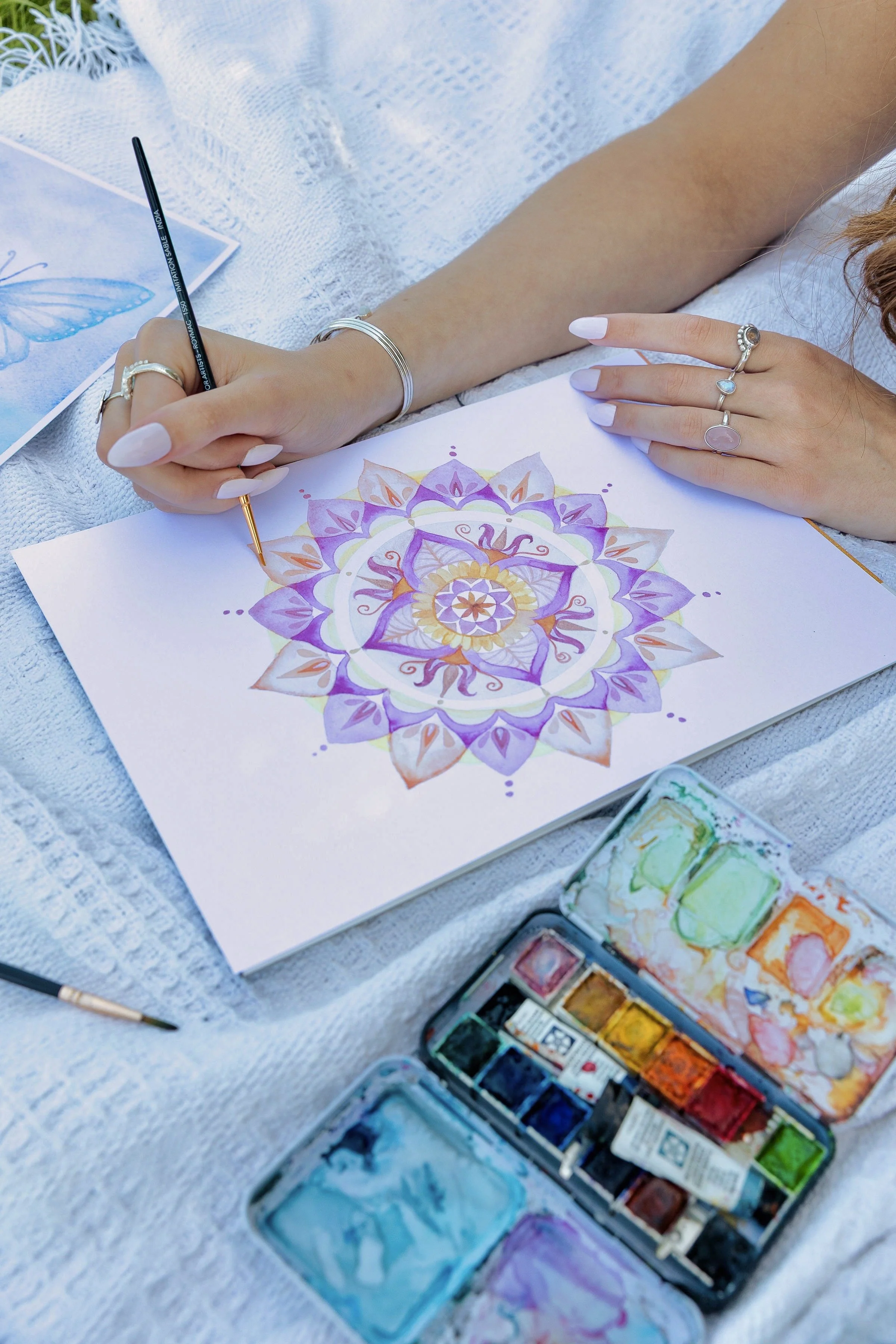 A person is creating a colorful mandala on paper with watercolors, surrounded by watercolor paint sets and brushes.