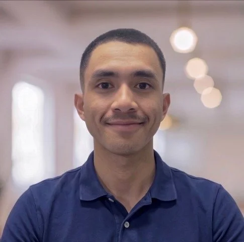 A man with short dark hair and a slight smile, wearing a navy blue collared shirt, standing indoors with blurred warm lighting in the background.