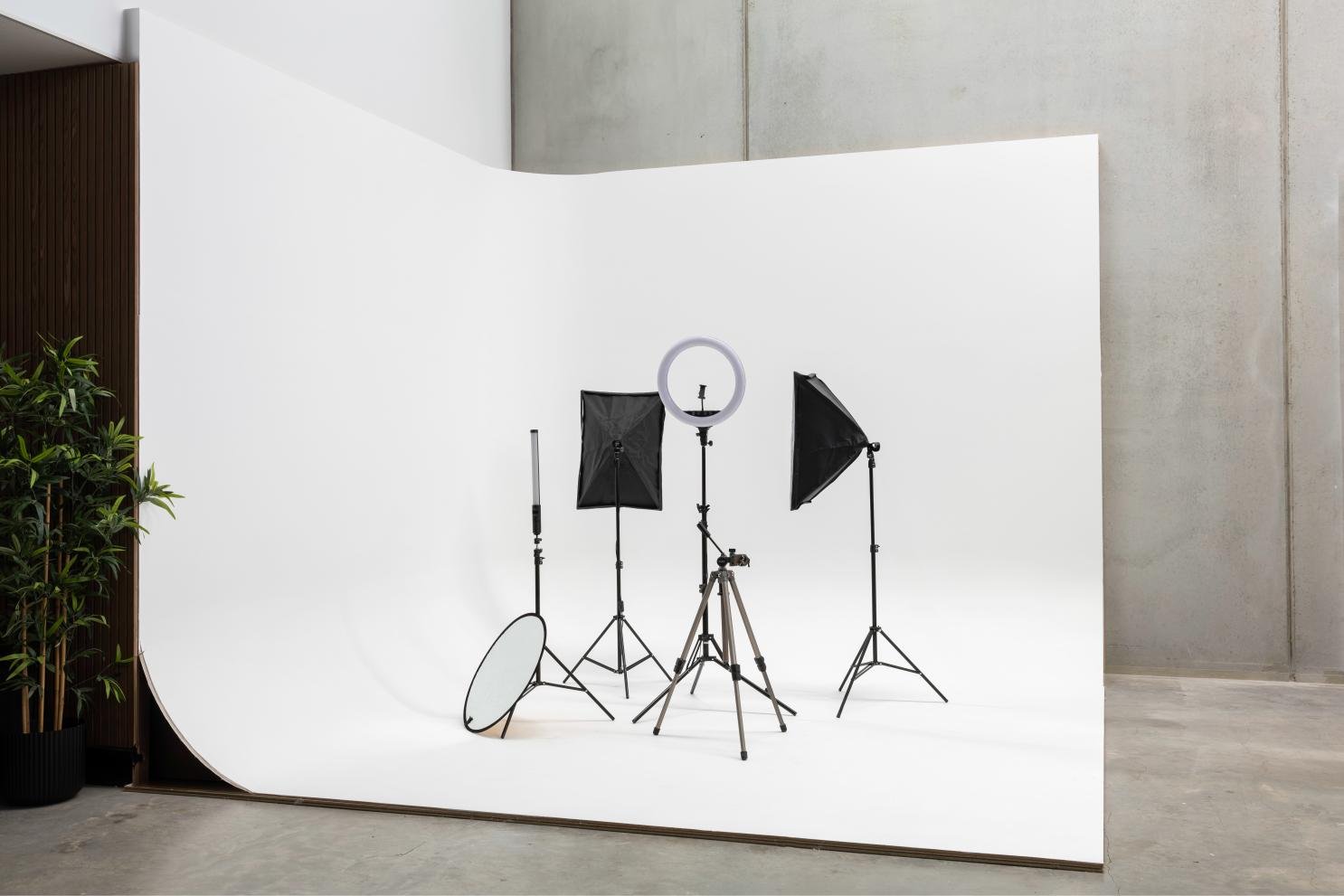 Photography studio setup with white backdrop, ring light on tripod, softboxes, reflector, and potted plant in the corner..jpg