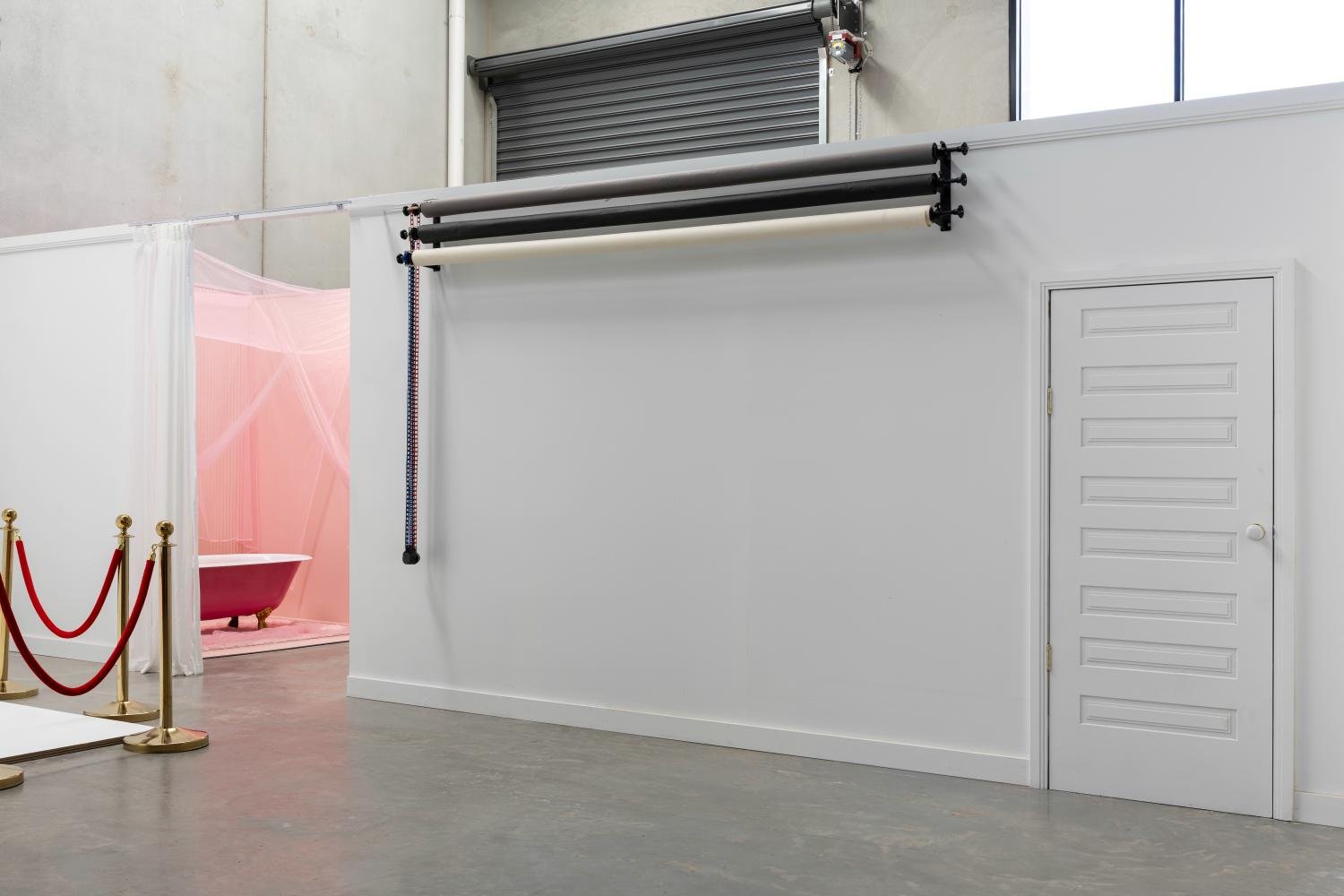 Interior of a photography studio with a white wall, several hanging backdrops, a closed door, and a visible pink bathtub surrounded by sheer curtains in an adjacent room..jpg
