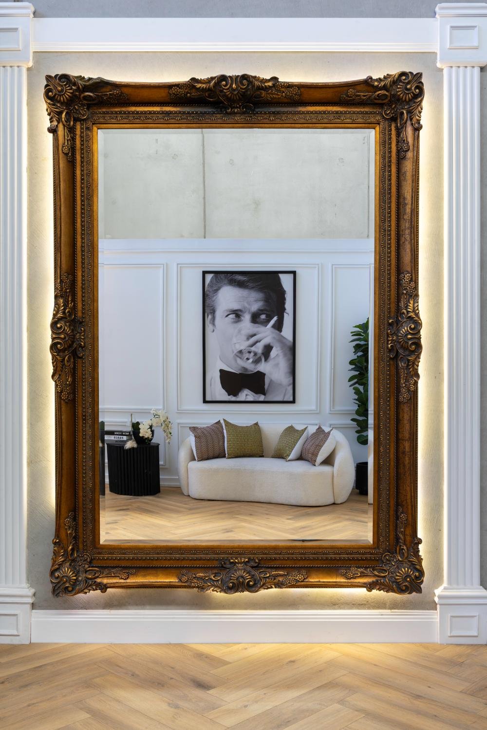 A large ornate mirror with a gilt frame reflects a modern living room featuring a white couch with decorative pillows. Behind the couch is a black-and-white portrait of a man, with paneled walls in th.jpg
