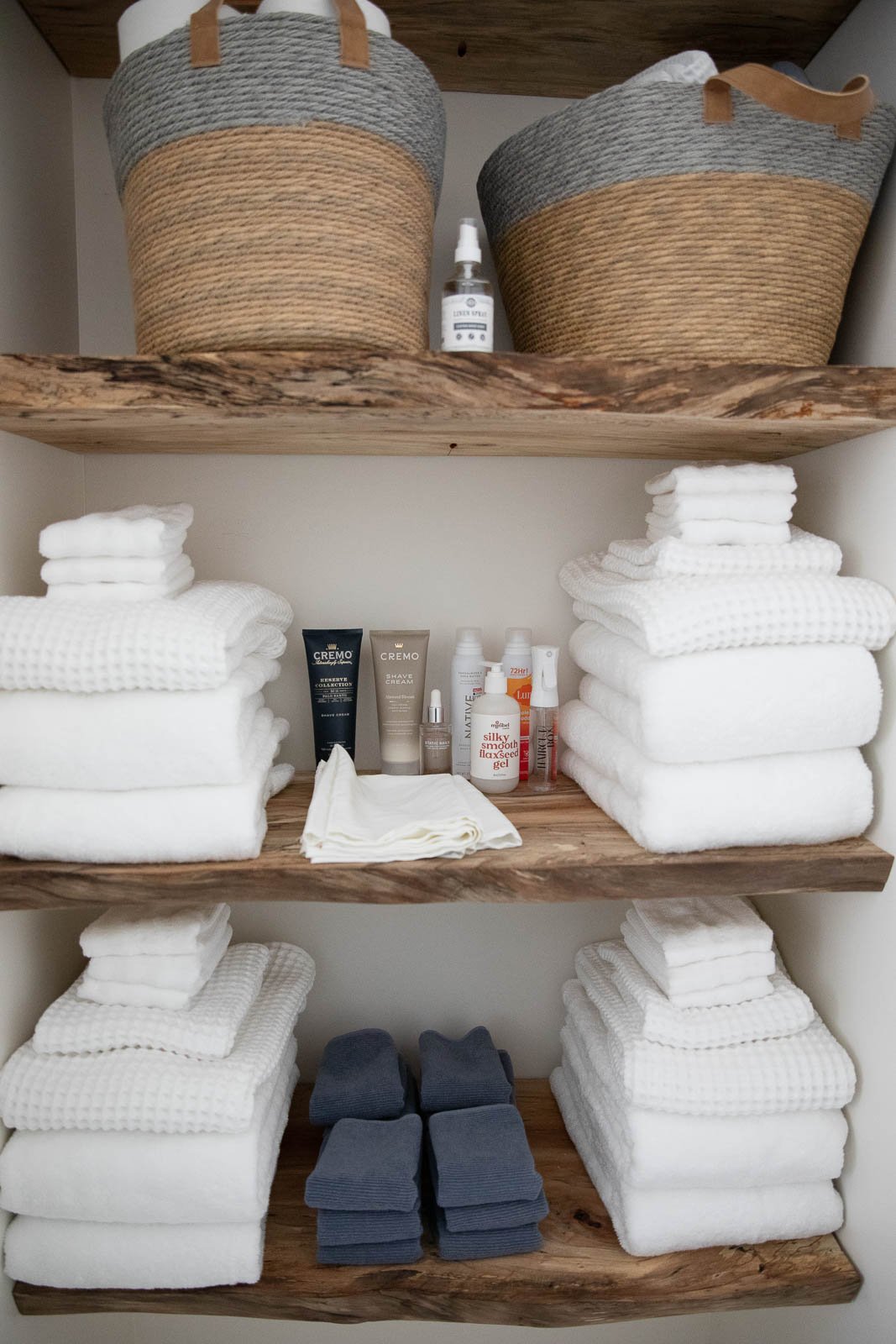 Wooden shelves with neatly folded white towels and dark gray washcloths, skincare and grooming products, and two woven baskets with handles.