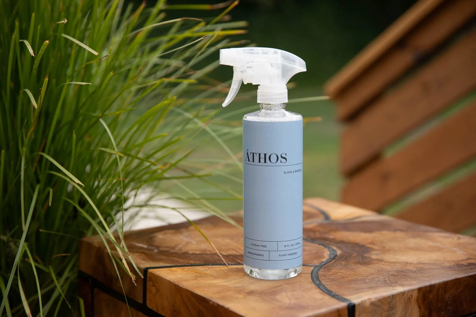 A spray bottle labeled 'ATHOS' on a wooden surface outdoors, with green plants to the left and a brick structure partially visible on the right.