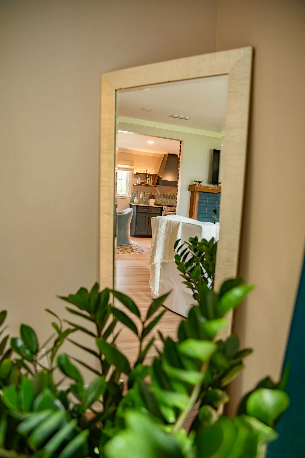 A mirror reflects a view of a cozy living room with a white couch, a fireplace, and a kitchen area with dark cabinets, a stove, and shelves with wine glasses, all seen through a mirror framed in light wood, with green plants in the foreground.