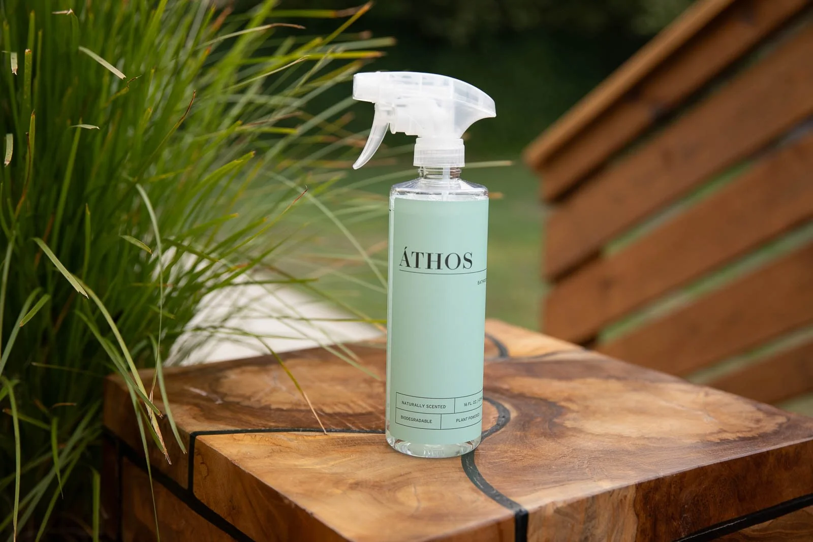 A spray bottle labeled 'ÁTHOS' sitting on a wooden surface outdoors, with green foliage in the background.