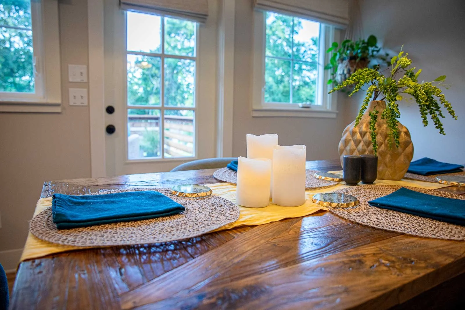 A wooden dining table set with four blue napkins, woven placemats, and a yellow table runner. In the center, there are three white pillar candles. On one side, there is a large geometric vase with greenery and flowers, and two small black cups. The b