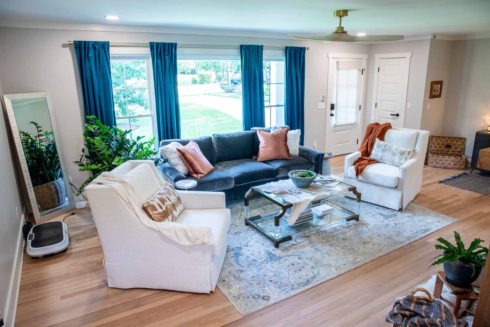 Living room with a large window, blue curtains, a dark sofa with pink pillows, white armchairs, a glass coffee table, a decorative rug, and various houseplants.