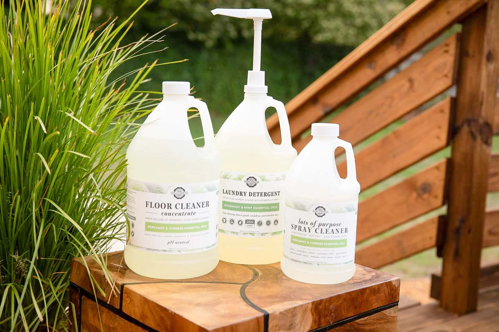 Three bottles of cleaning products placed on a wooden surface outdoors, with greenery and a wooden structure in the background. The bottles are labeled as floor cleaner, laundry detergent, and spray cleaner, featuring essential oils in their ingredie