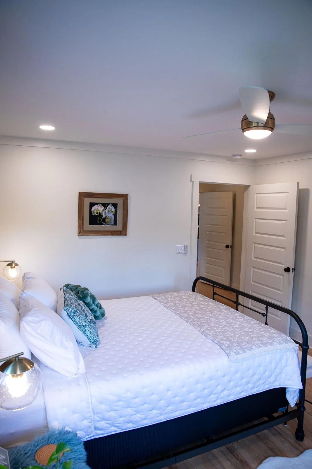 A bedroom with a white bed against a white wall, a framed floral picture hanging above, a bedside table with a glass lamp, and a ceiling fan with a light. There are two open closet doors.