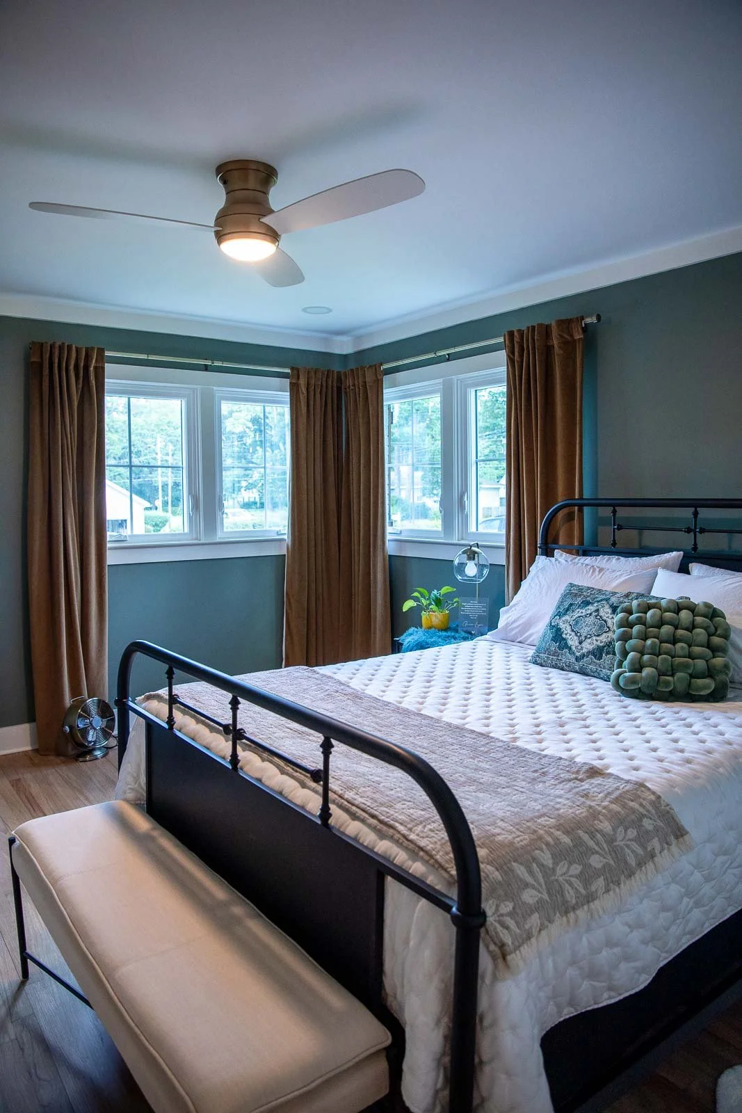 A bedroom with a black metal bed frame, white and patterned pillows, and a beige quilt. There are three windows with brown curtains, a beige bench at the foot of the bed, and a ceiling fan with light fixtures. A small side table holds a plant and a d